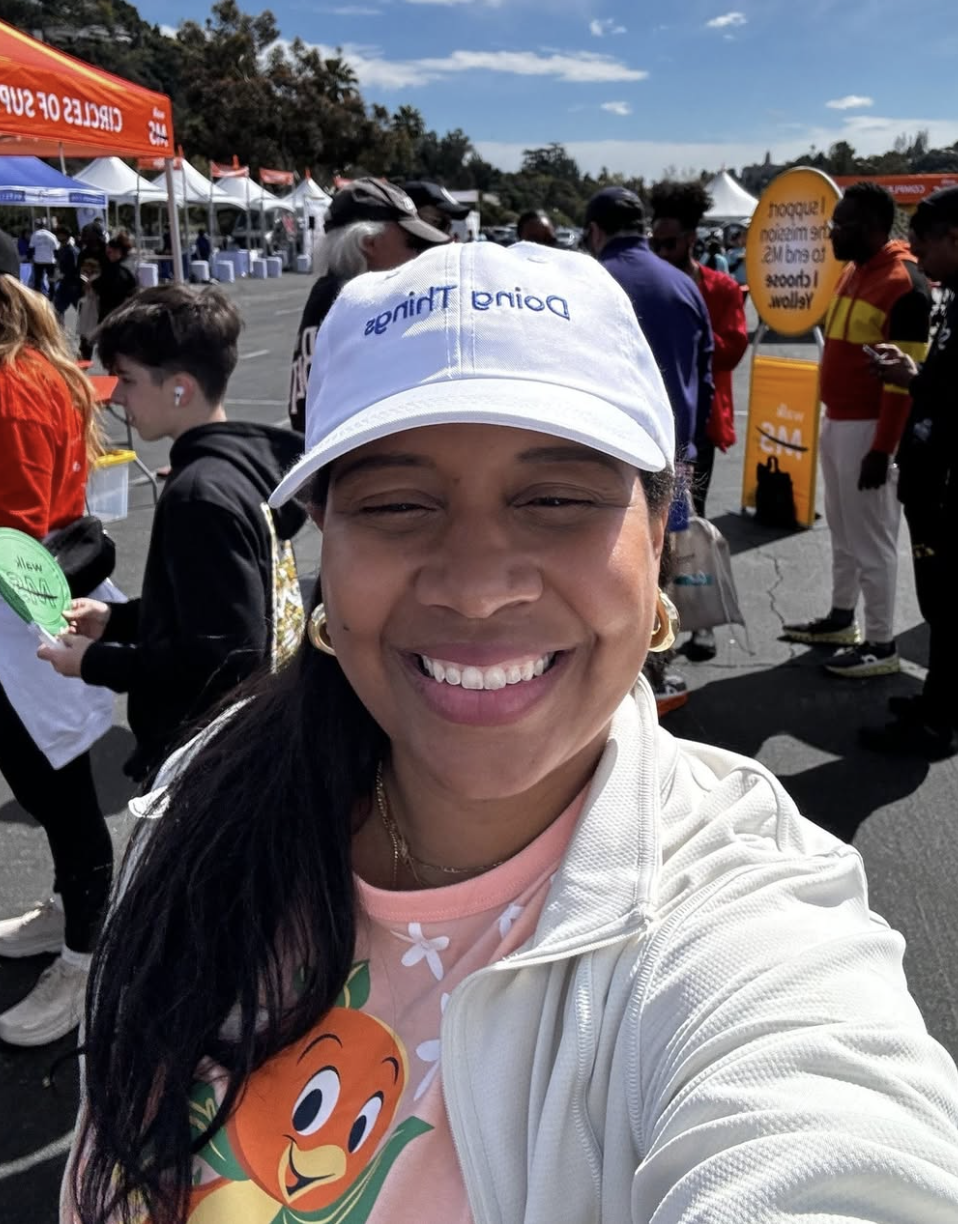 Smiling woman taking selfie at an outdoor event with tents and many people in the background.