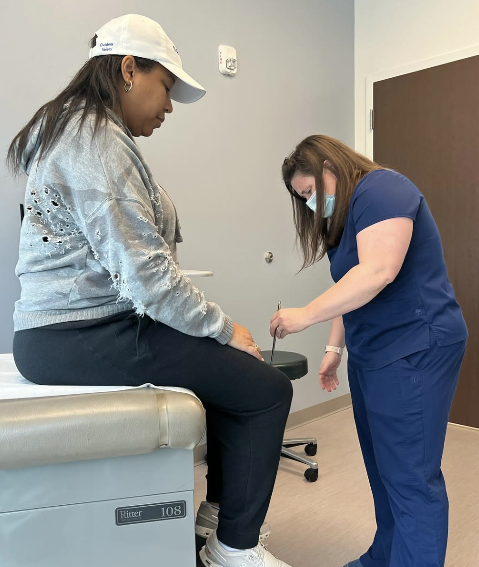 A healthcare professional in blue scrubs, wearing a mask and glasses, is preparing check on Marti Hine's progress in her ongoing battle with Multiple Sclerosis, who is seated on a medical examination table, in a clinical setting.