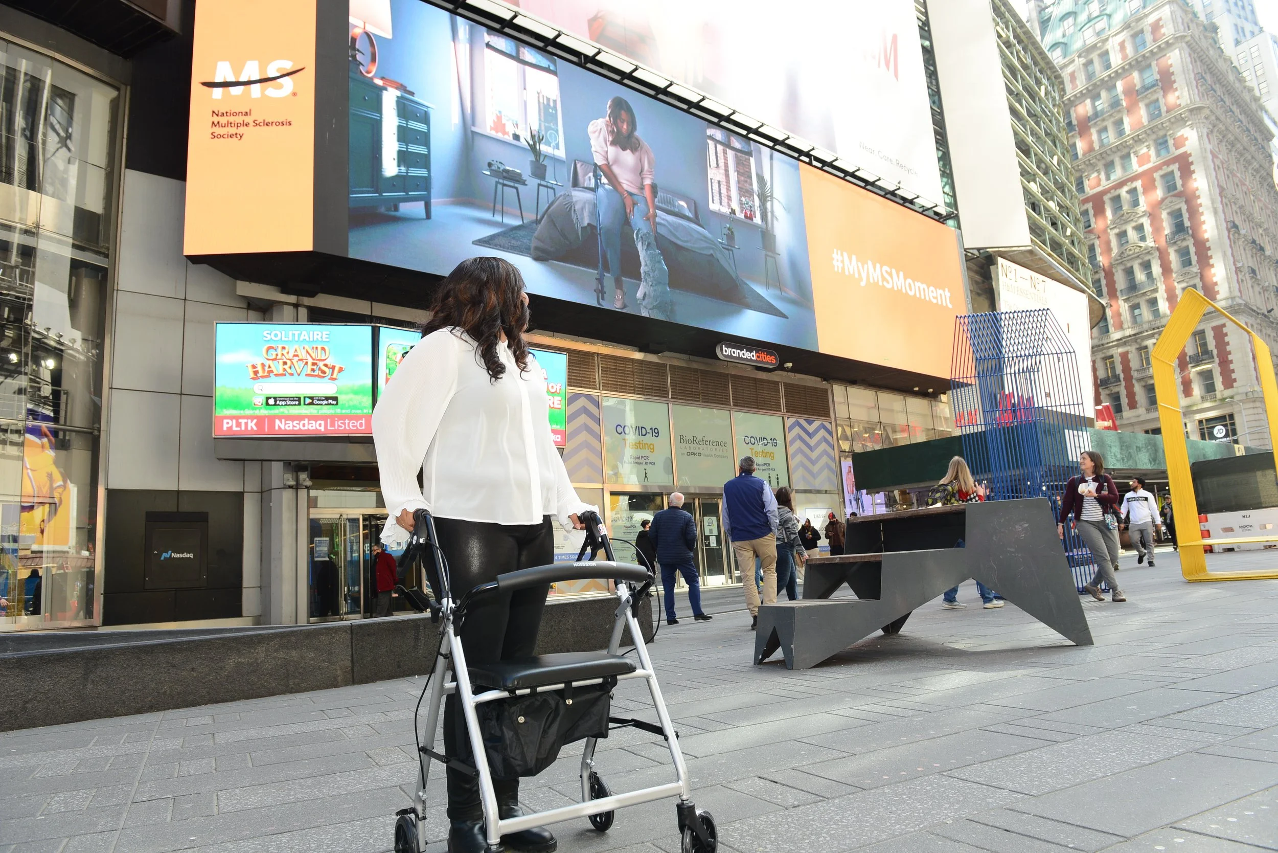 Marti with dark hair wearing a white blouse and black pants, using a walker, stands on a busy city street in front of large digital billboards. Other pedestrians walk by in the background.