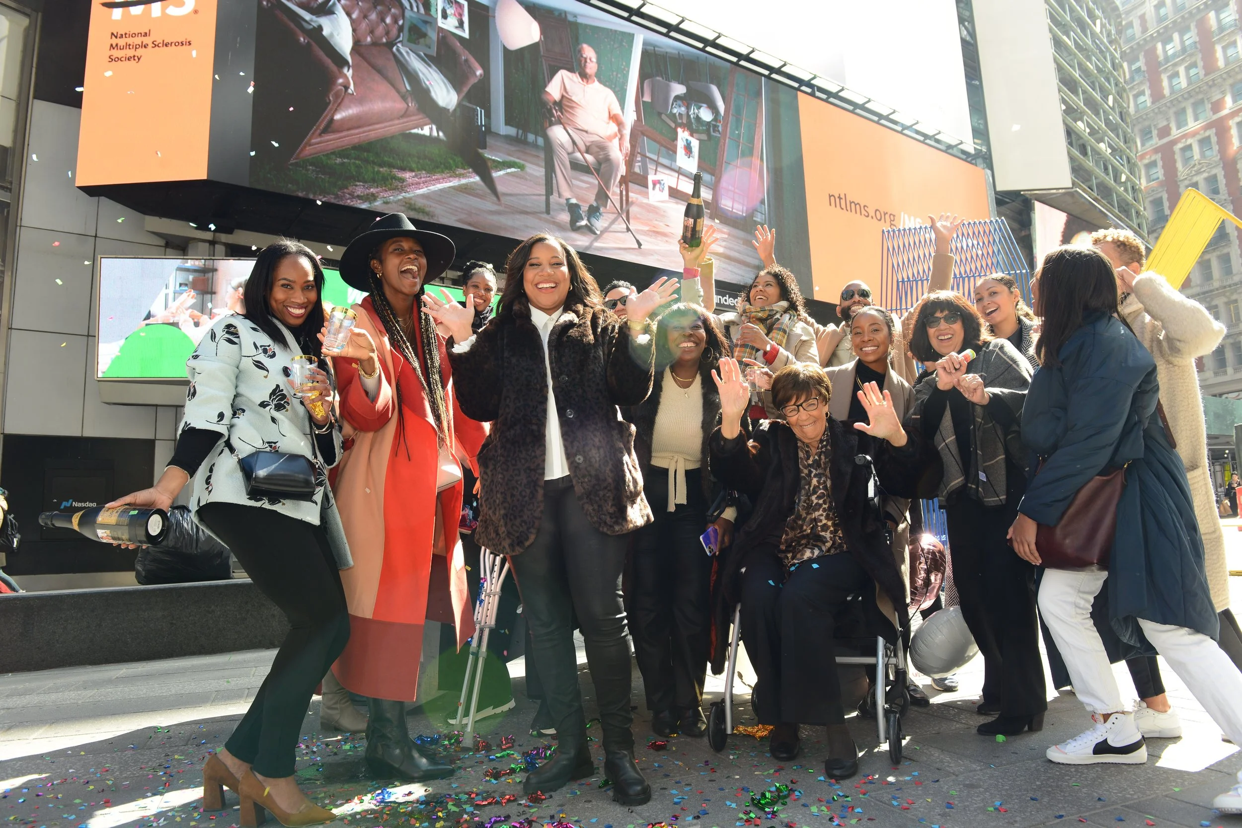 A diverse group of women celebrating outdoors in Times Square, New York City, with confetti on the ground and large screens in the background.