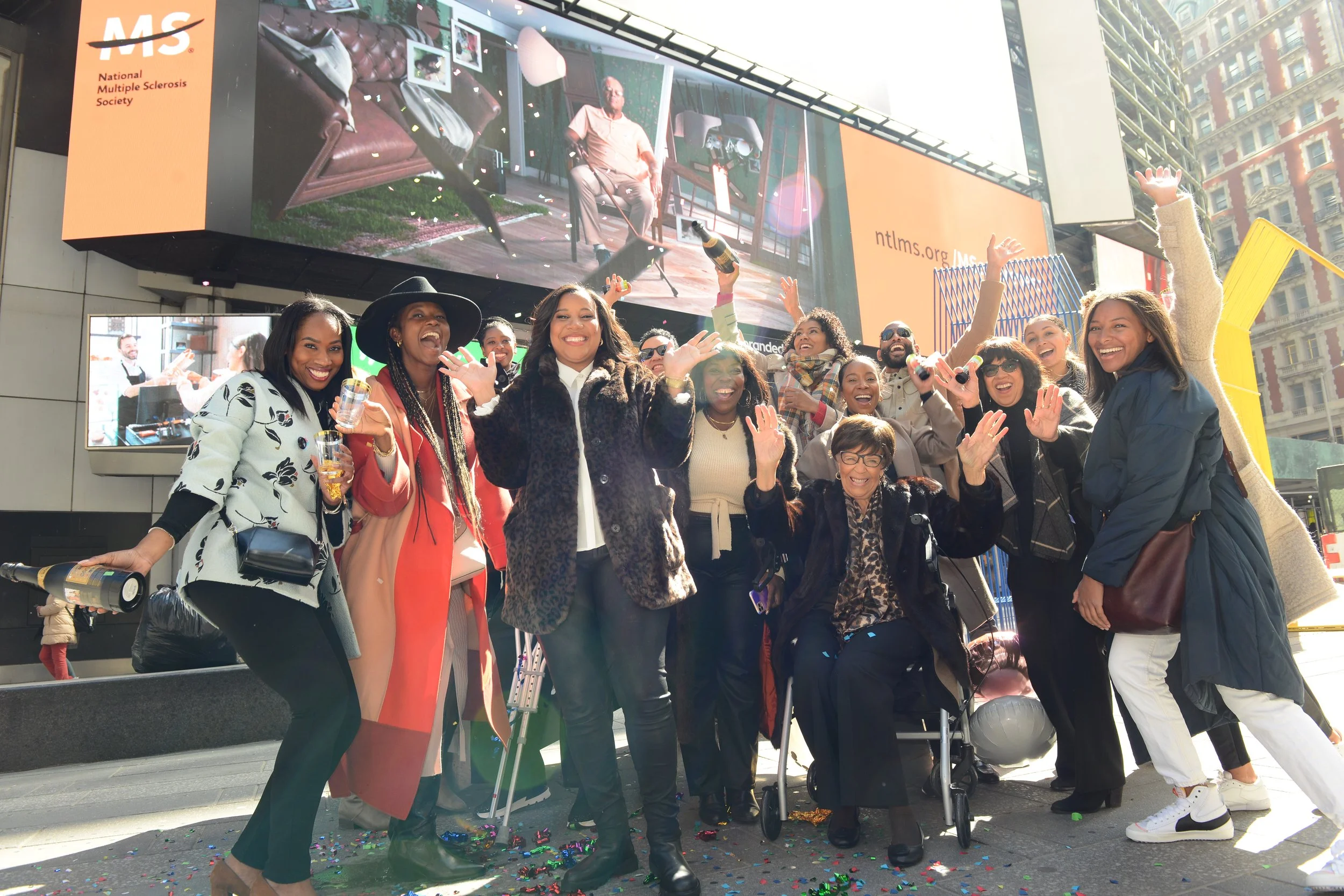 Group of people celebrating outdoors in front of digital billboards, with some holding drinks, waving, and smiling.