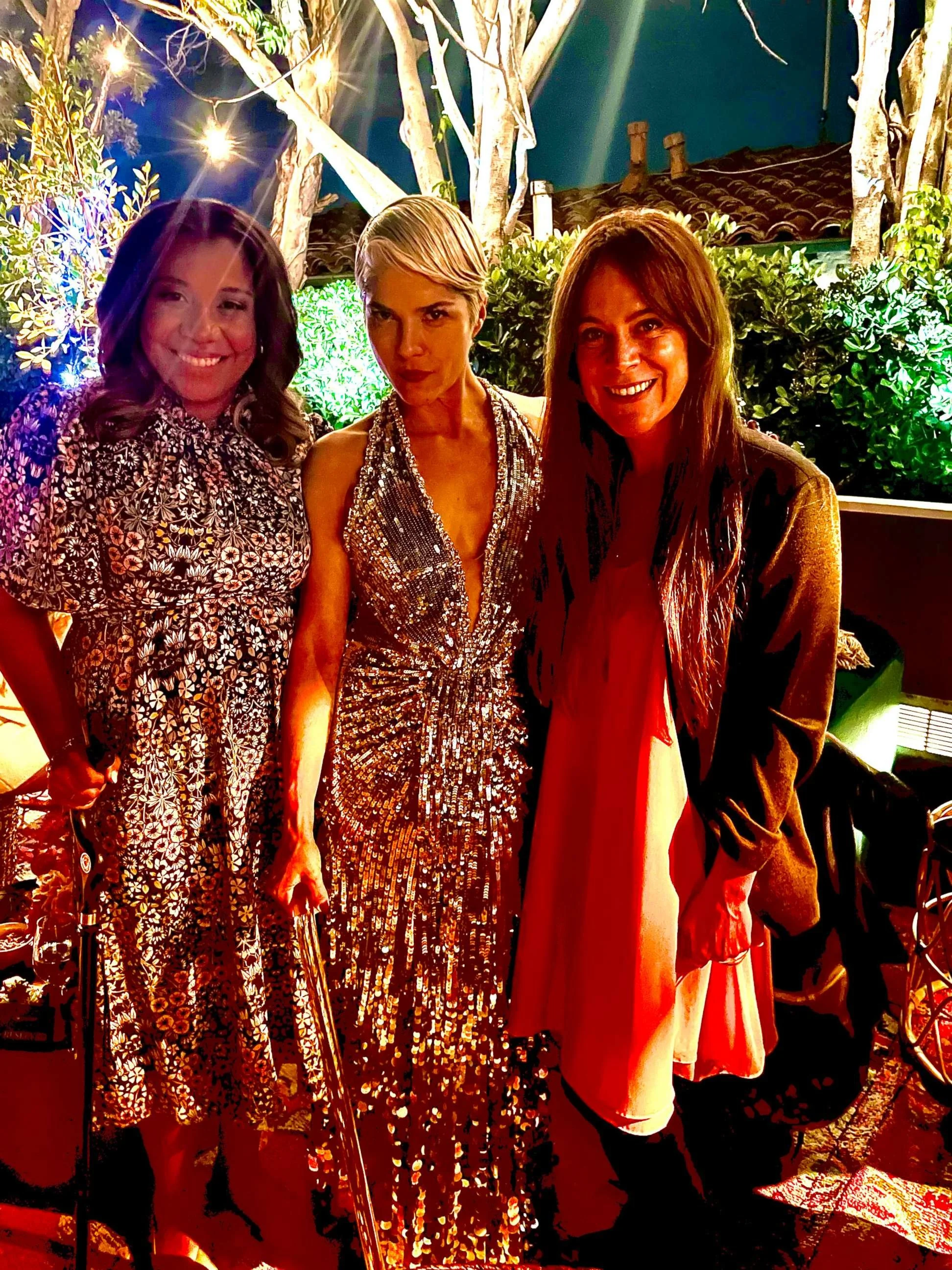 Three women dressed in elegant evening attire standing outdoors at night, with trees, bushes, and string lights in the background.
