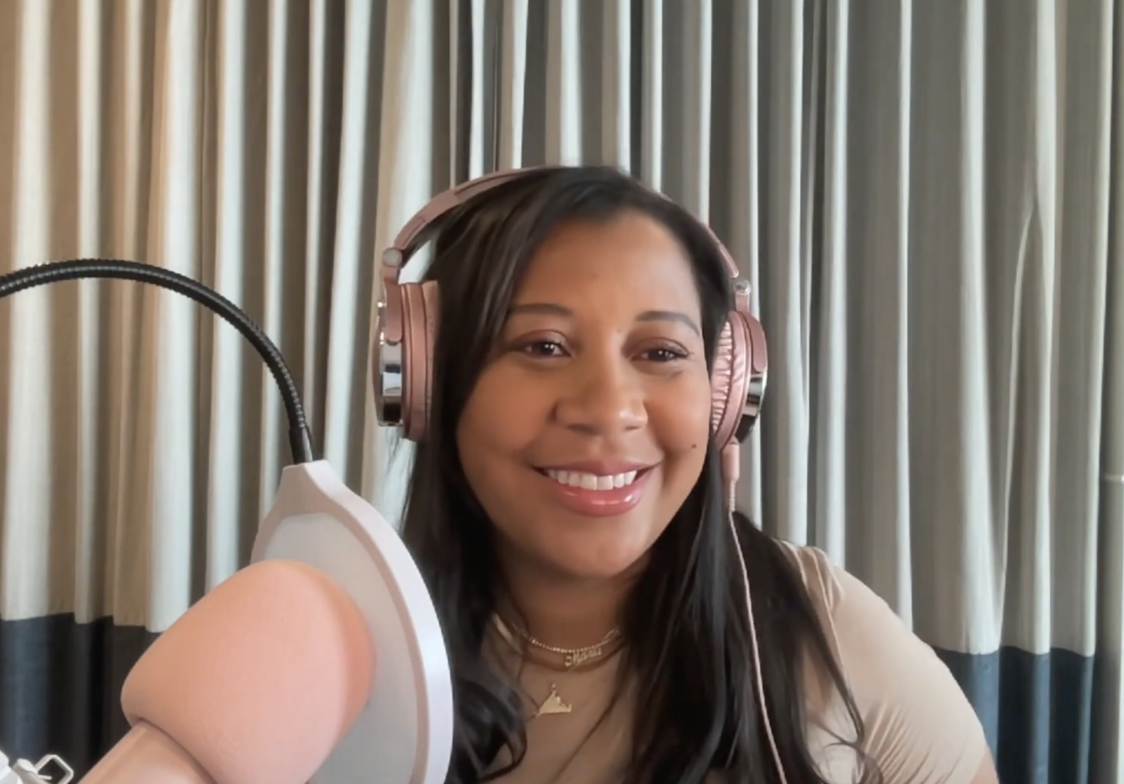 Marti Hines, a woman with long dark hair wearing pink headphones and a beige top, smiling in front of a blue curtain, with a large pink microphone positioned near her face.