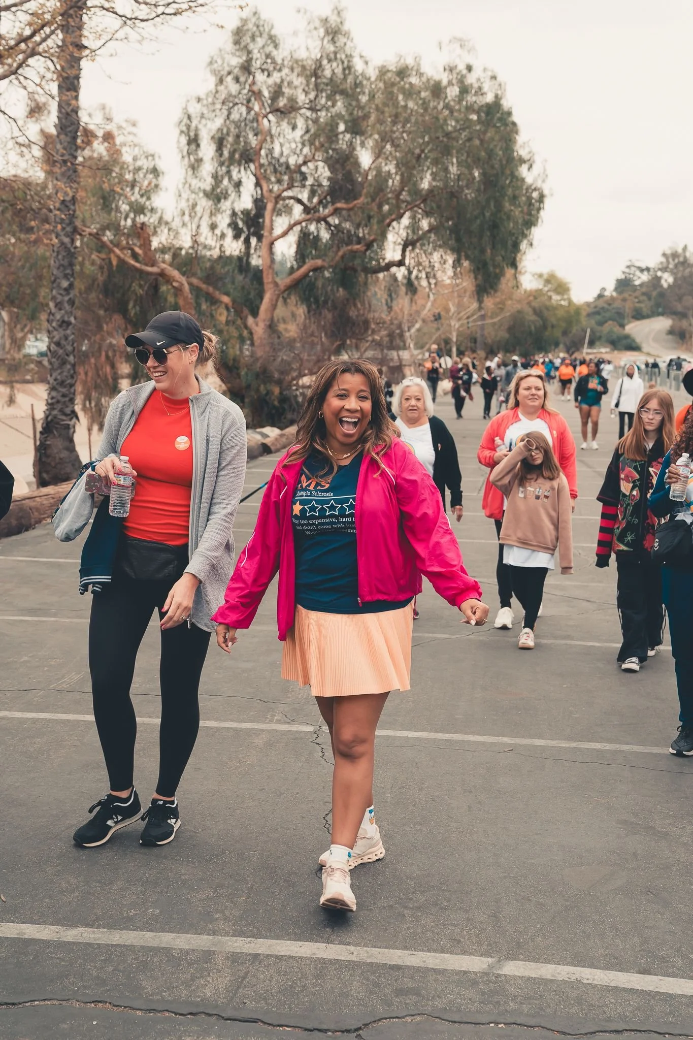 Group of women, including Marti Hines, walking outdoors in a park-like area, smiling and enjoying themselves, with trees and a road in the background. The event is for Multiple Sclerosis Awareness.