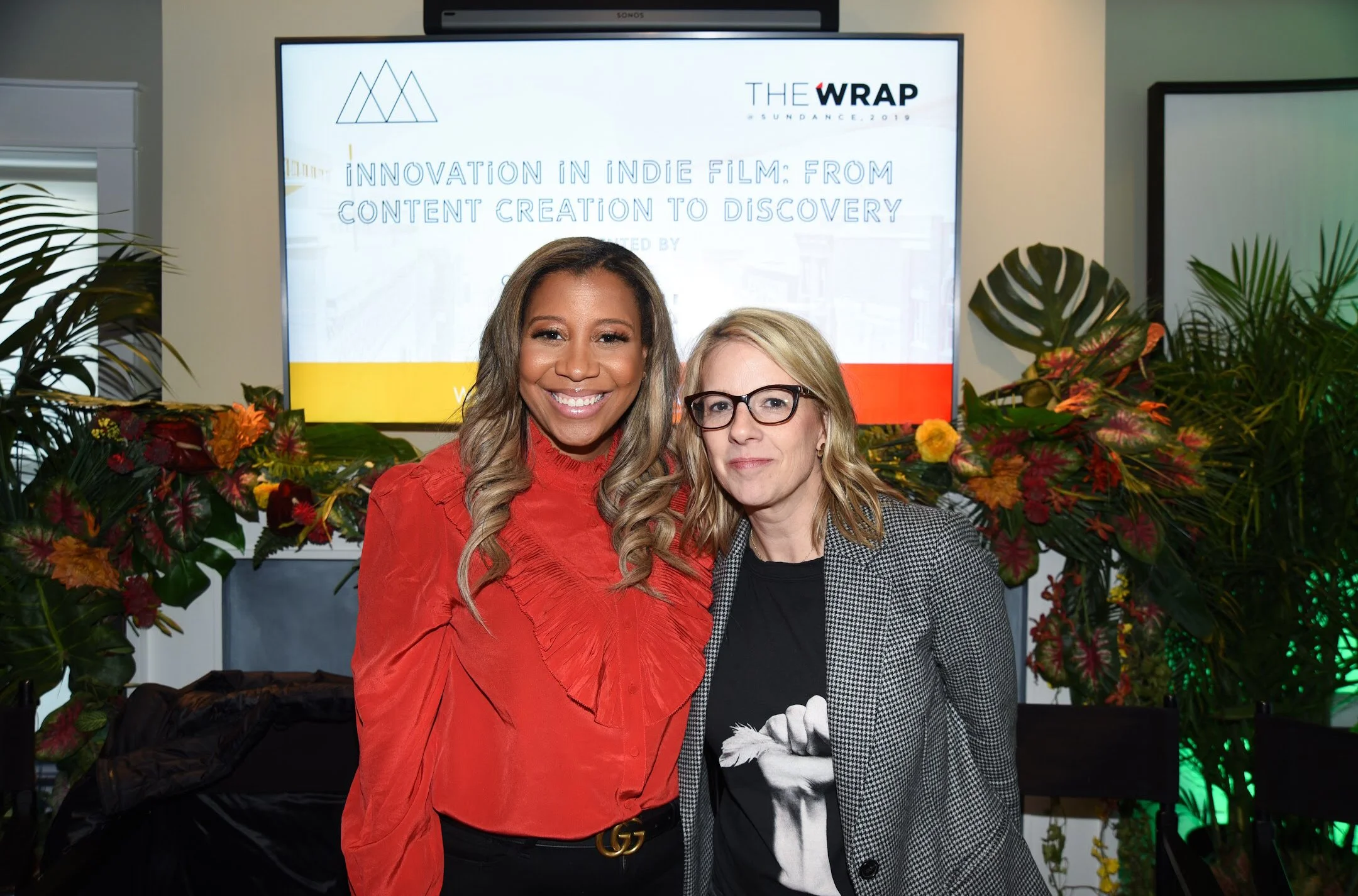 Marti Hines and friend standing together at an indoor event, smiling at the camera. Behind them is a large screen with a presentation title that reads 'Innovation in Indie Film: From Content Creation to Discovery,' accompanied by decorative plants.