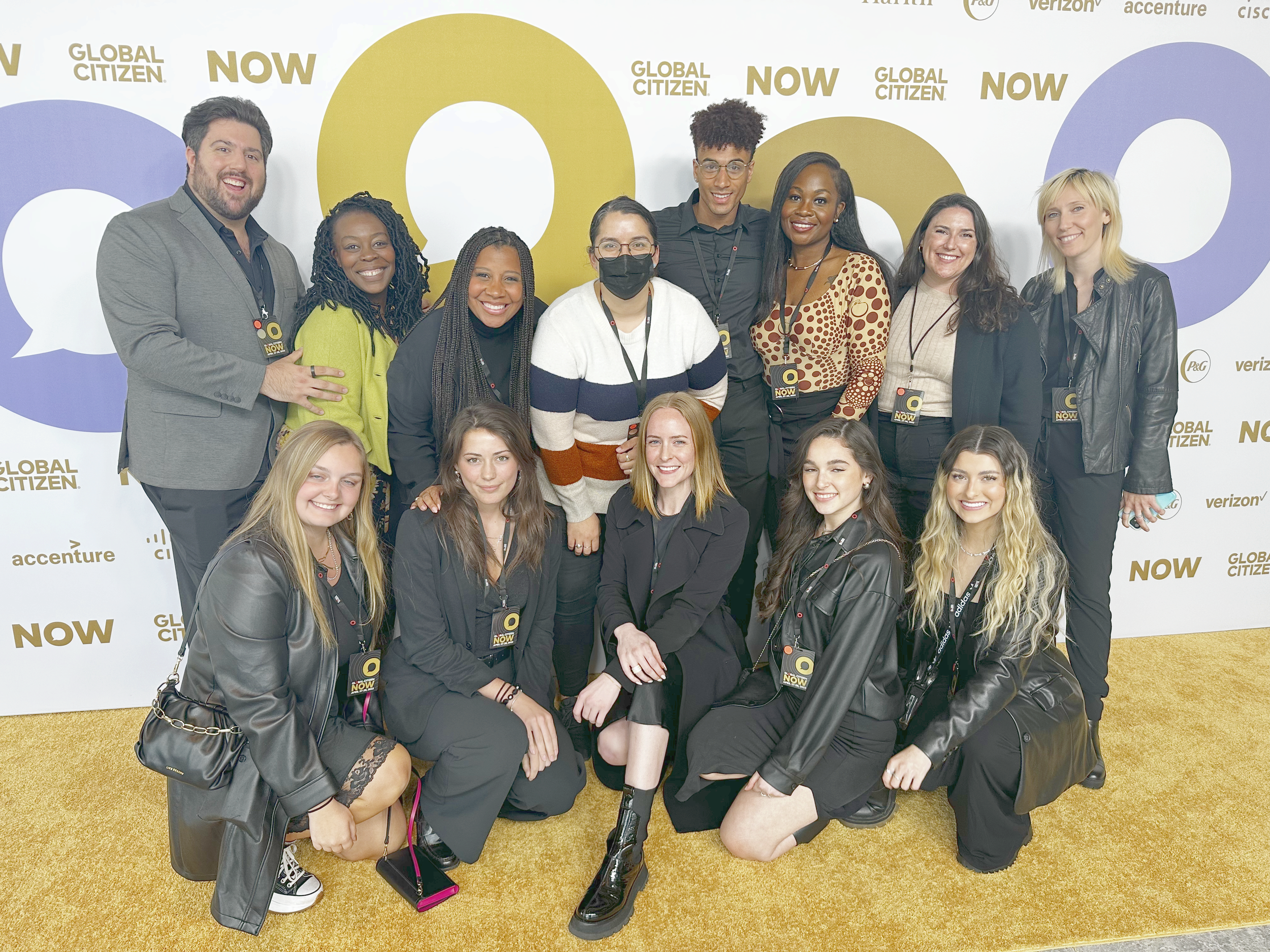 Group of fifteen diverse young adults posing at a Global Citizen event, with a backdrop featuring logos of Global Citizen, NOW, Verizon, and other sponsors. They are smiling and dressed in casual and business casual attire, some wearing event badges.