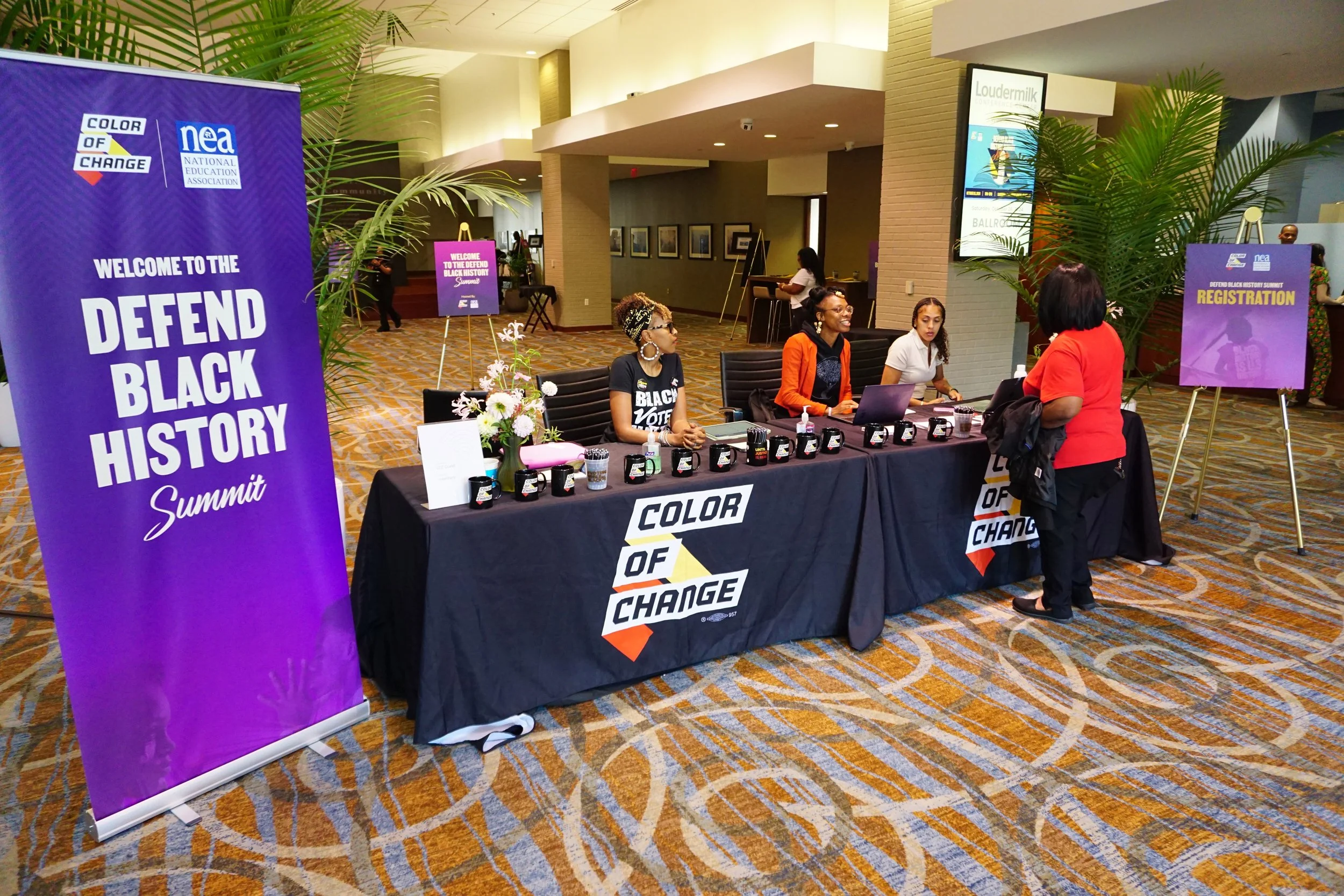 Registration desk at the 'Defend Black History Summit' with women sitting behind the table, a woman standing in front talking to the staff, and banners promoting the event.