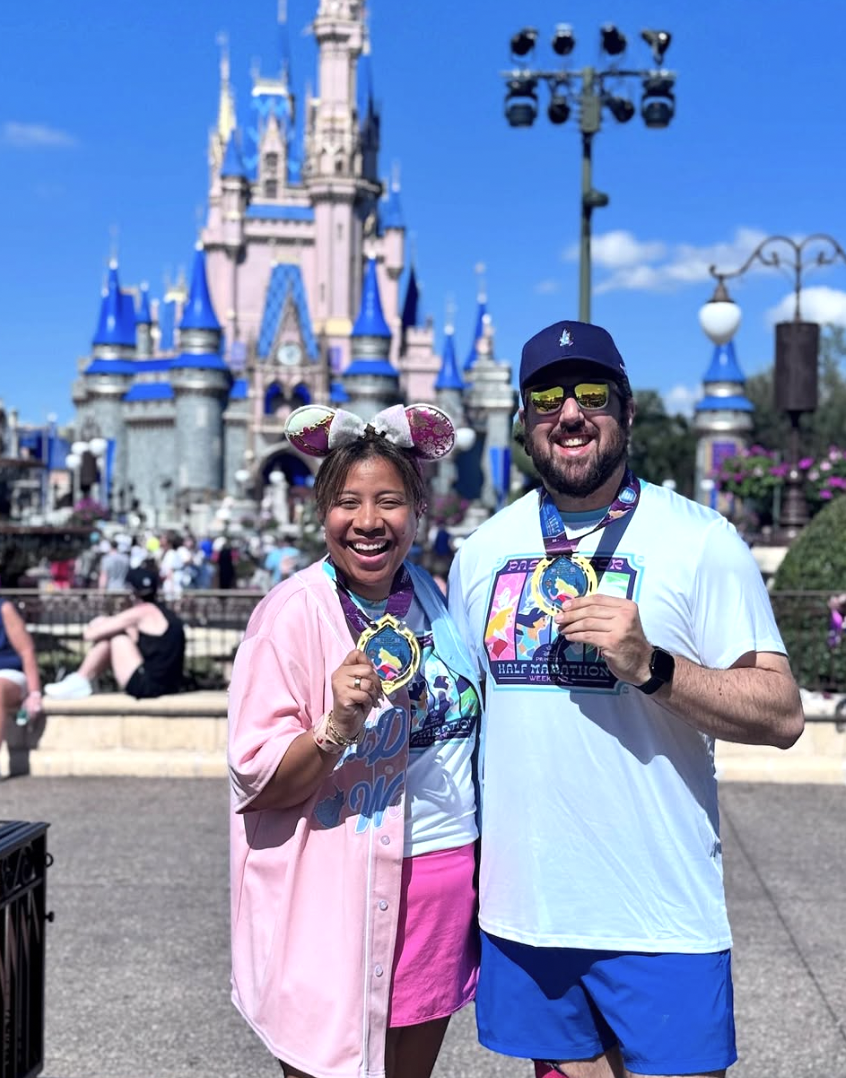 Two smiling people standing in front of Cinderella's Castle at Disney theme park, holding medals from a race event. The woman wears Mickey Mouse ears and a pink outfit, while the man wears sunglasses, a cap, and a white t-shirt.