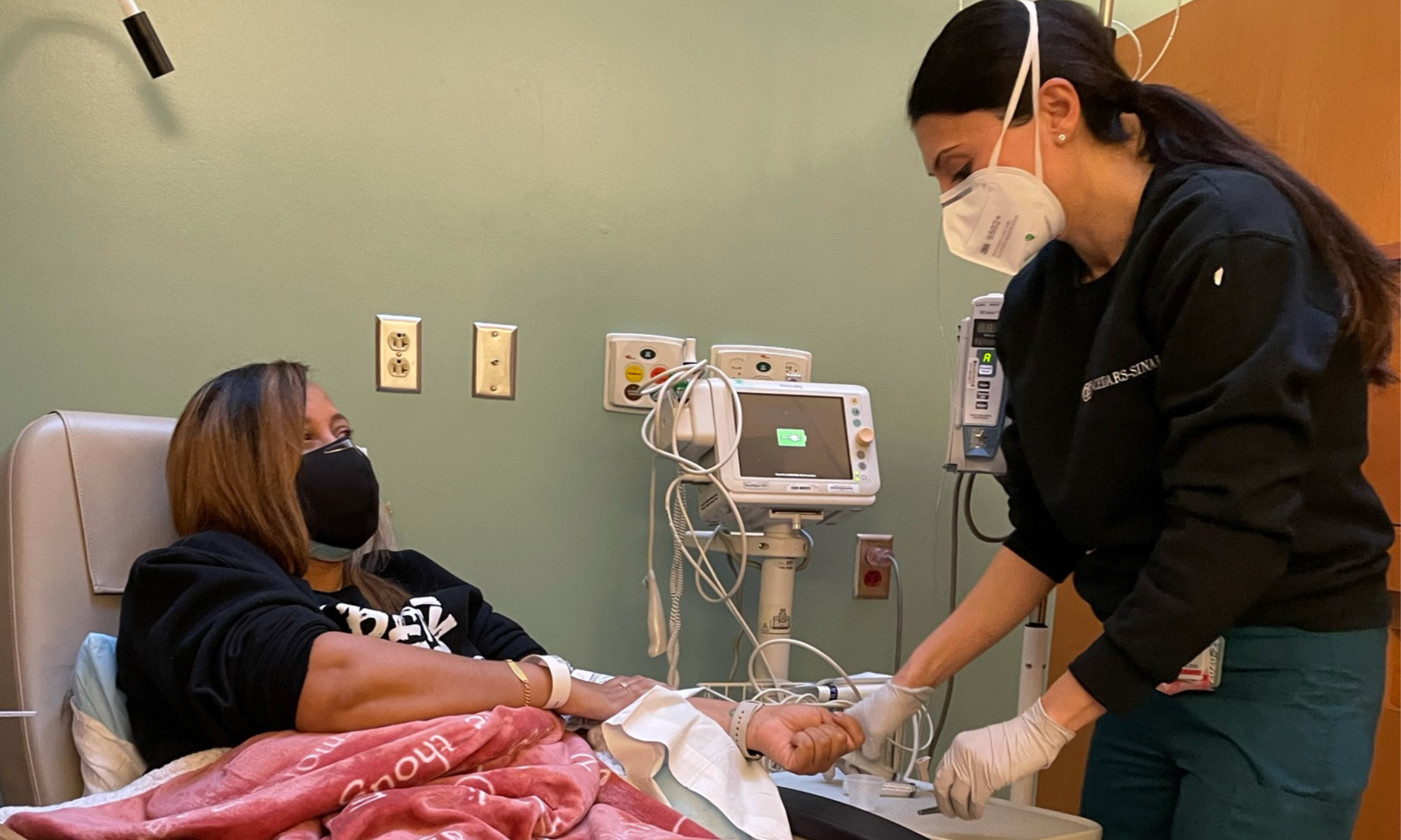 Marti Hones with a black face mask is lying in a hospital bed connected to medical equipment as she receives treatment for Multiple Sclerosis. A healthcare worker wearing a face mask, gloves, and black scrubs is holding the Marti's hand.