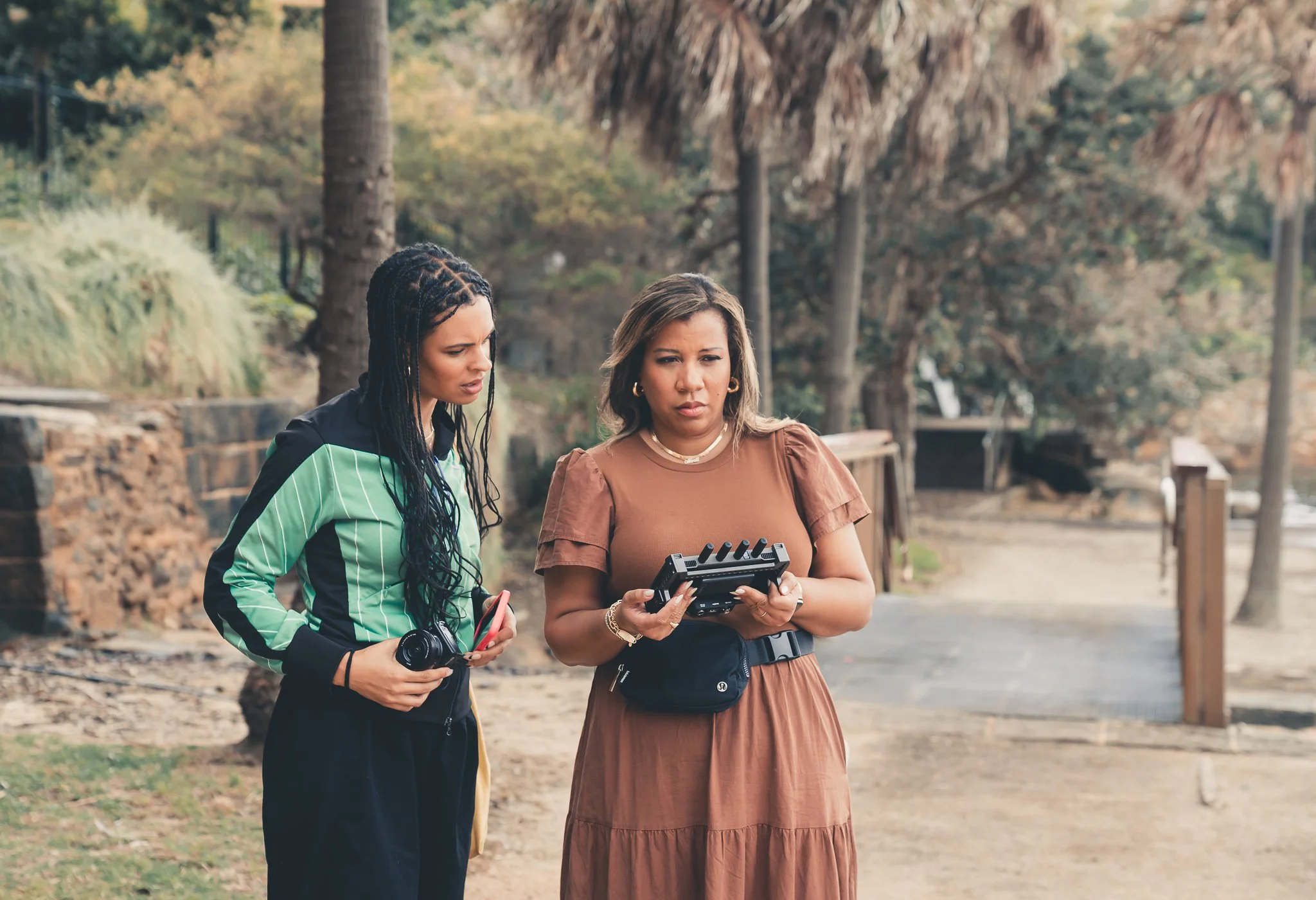 Marti Hines and female friend standing outdoors in a park-like setting, examining a camera and camera accessories, with trees and a pathway in the background.