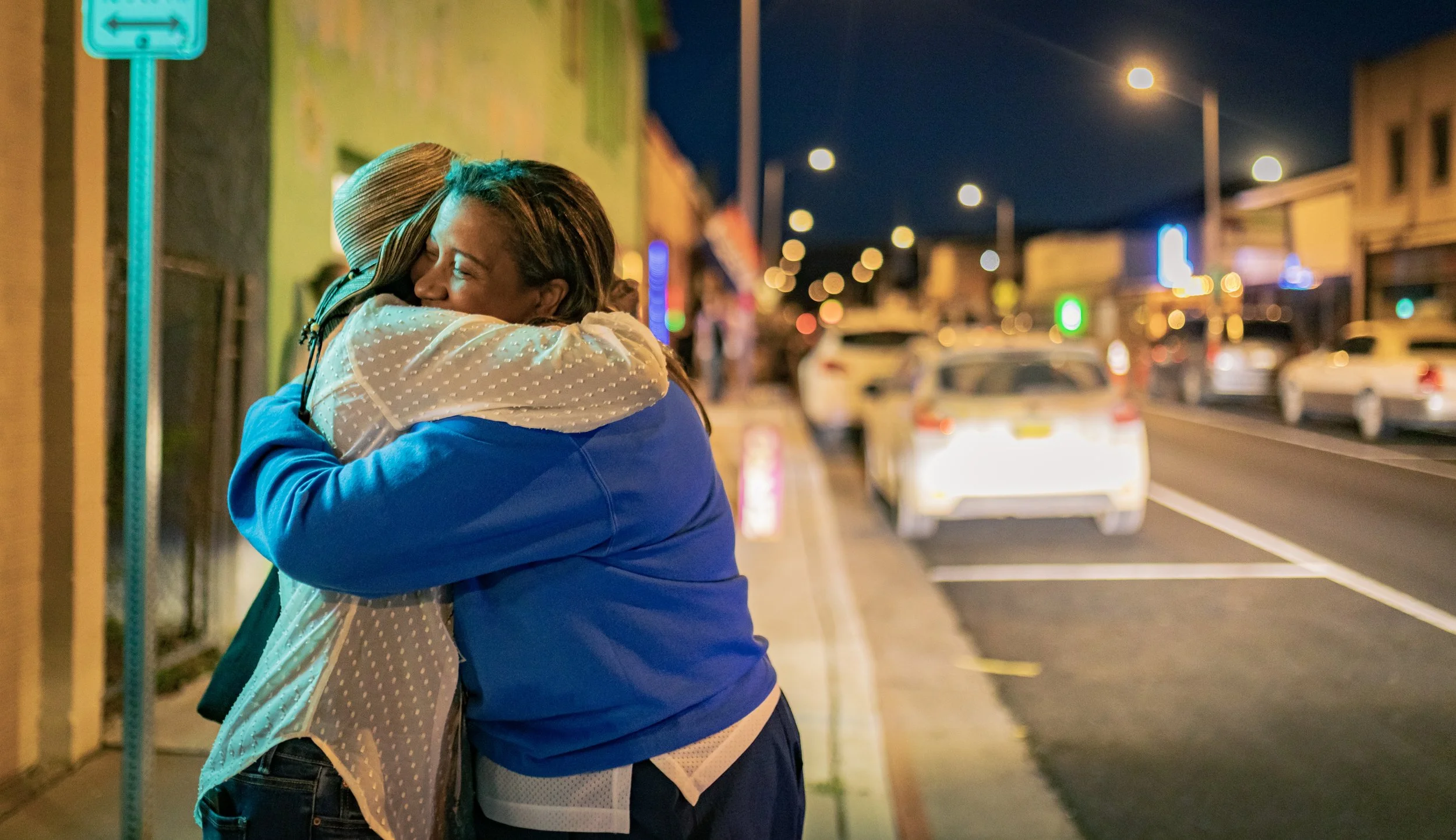 Two women, Marti and friend, hugging on a city sidewalk at night, with cars parked along the street and blurred city lights in the background.