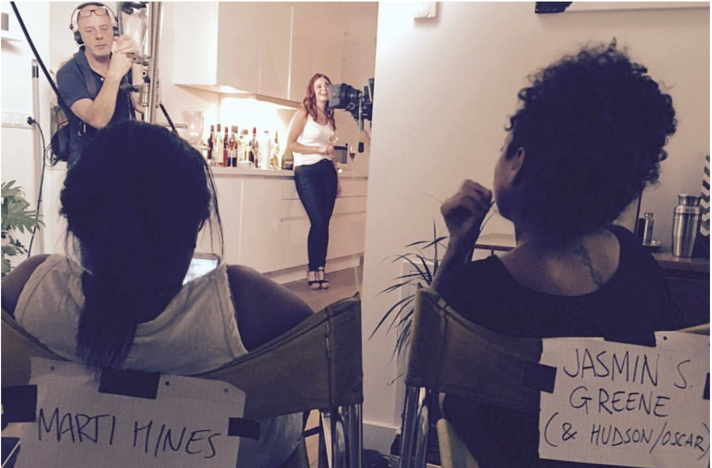 Behind-the-scenes photo of a film or photo shoot in a modern kitchen. A woman, Jasmine S. Greene, is standing in front of a camera, smiling, while two people are seated in the foreground with signs reading 'Marti Hines' and 'Jasmin S. Greene (& Hudso
