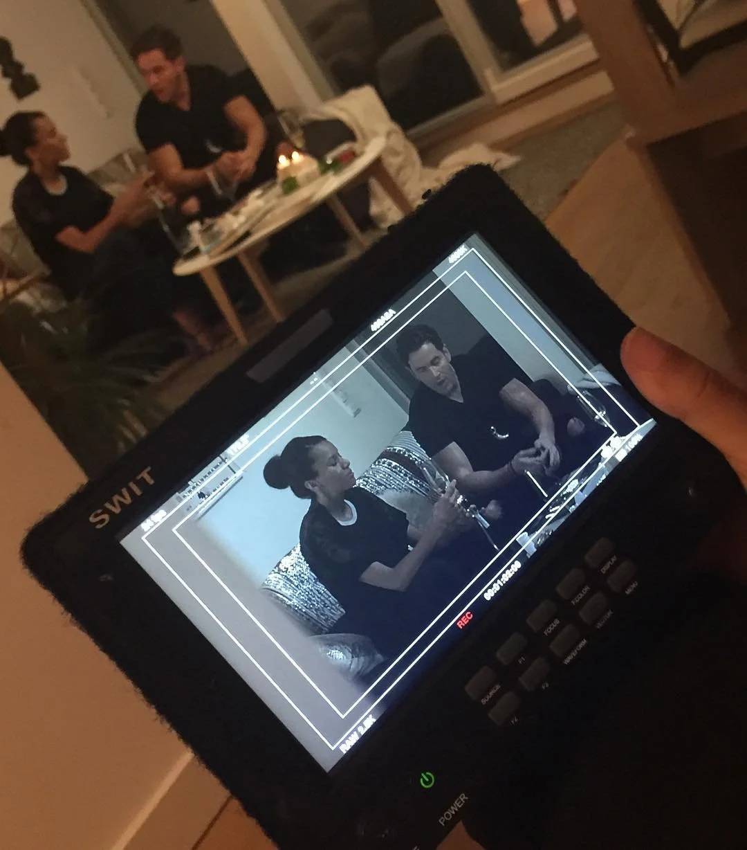 A camera screen showing a man and a woman seated on a patterned sofa, engaged in conversation, with the man wearing a black shirt and the woman holding a glass, while in the background, two people are sitting at a table with drinks in a living room.