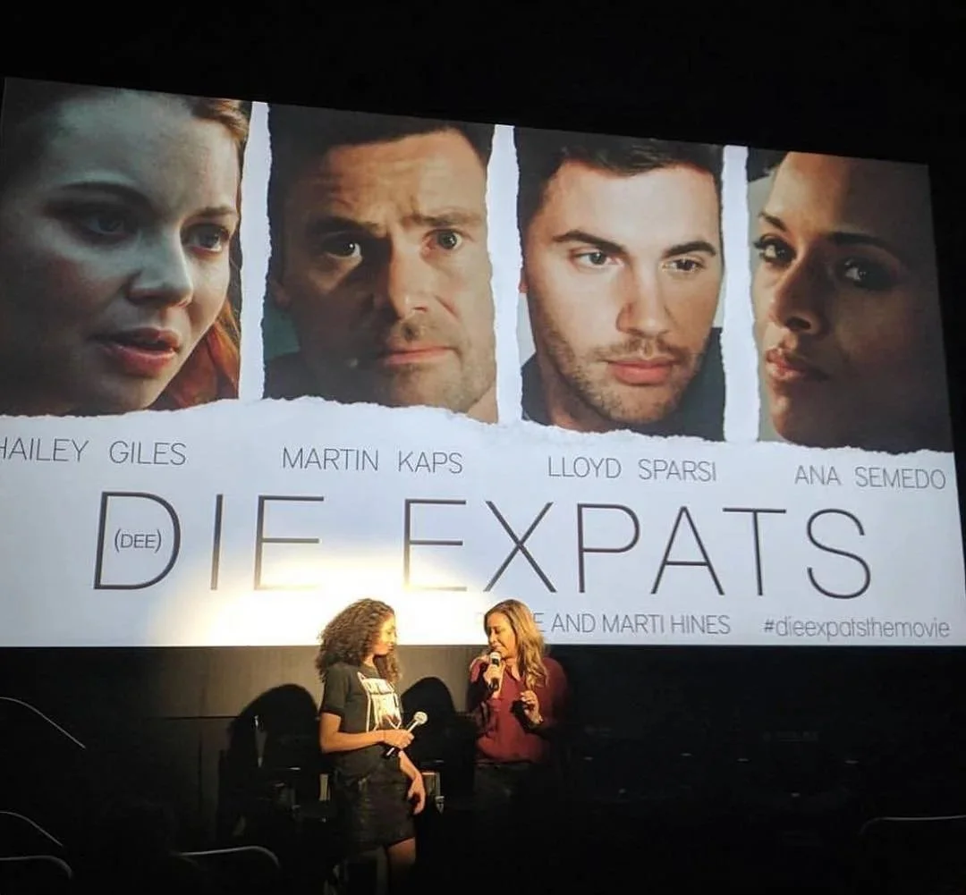 A movie poster titled 'Die Expats' featuring images of four actors: Hailey Giles, Martin Kaps, Lloyd Sparsi, and Ana Semedo. Two women are standing in front of the poster, hosting a discussion or presentation.