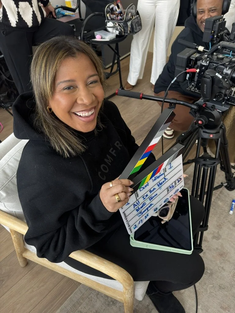 A woman smiling and holding a clapperboard and a tablet, sitting in a chair. Behind her are filming equipment and crew members on a film or TV set.