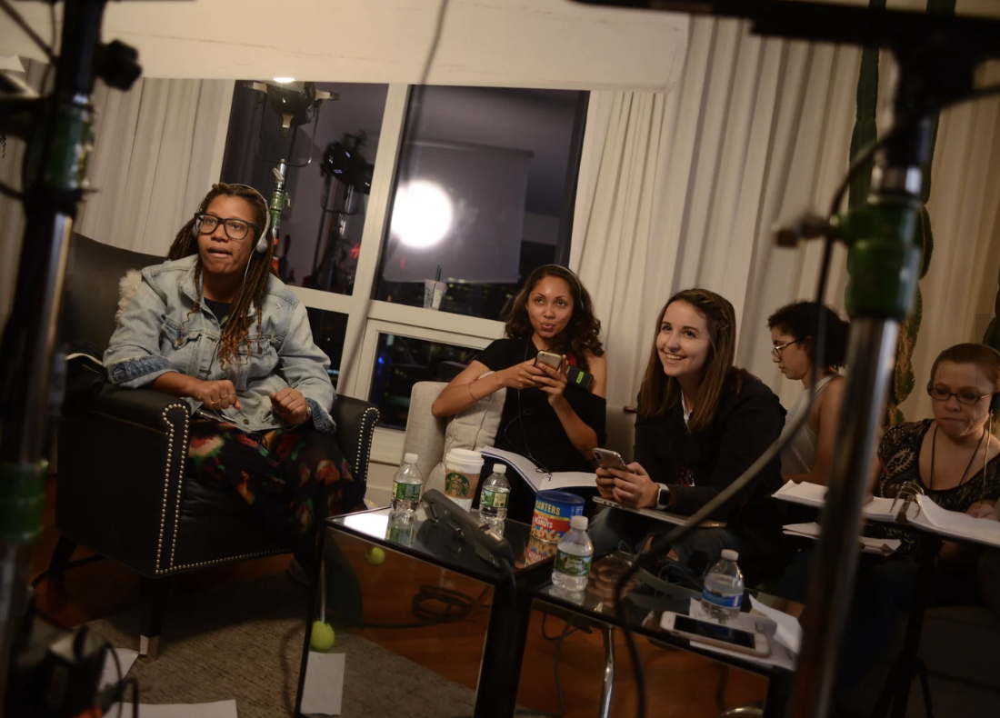 A group of five women sitting in a room, engaged in a discussion or meeting, with equipment and bottles of water on the table.