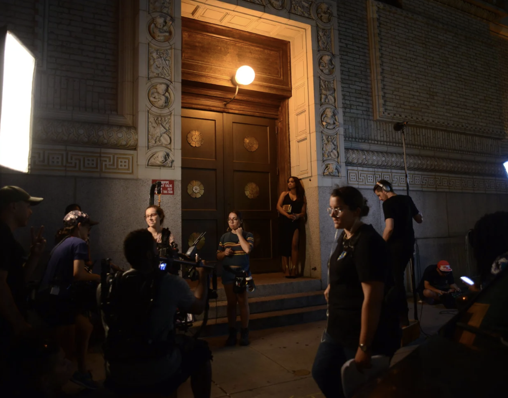 Film or video production crew setting up outside a building with ornate architecture, preparing for a shoot at night.