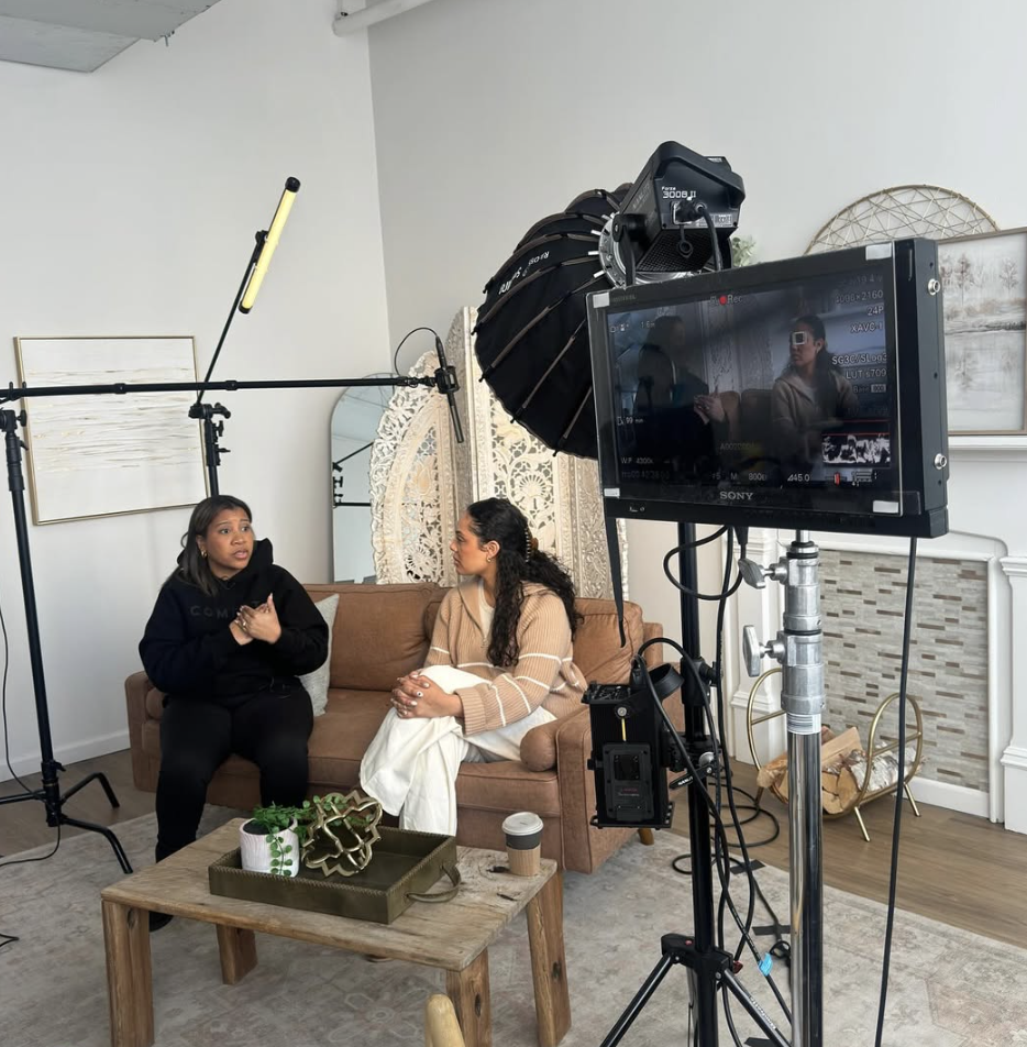 Two women sitting on a tan sofa engaged in conversation during a video recording session, with filming equipment including a camera on a stand, a large softbox, and a monitor visible in a well-decorated room.