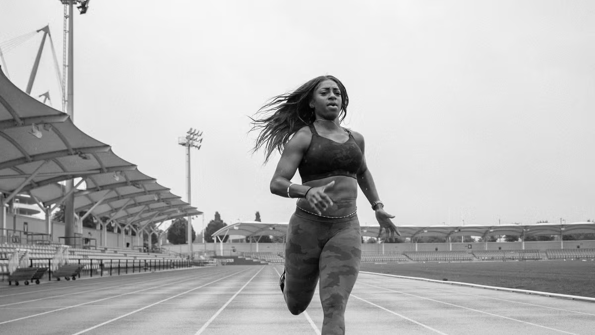 A woman running on a track at an outdoor stadium, wearing athletic clothing, with empty stands and a cloudy sky in the background.