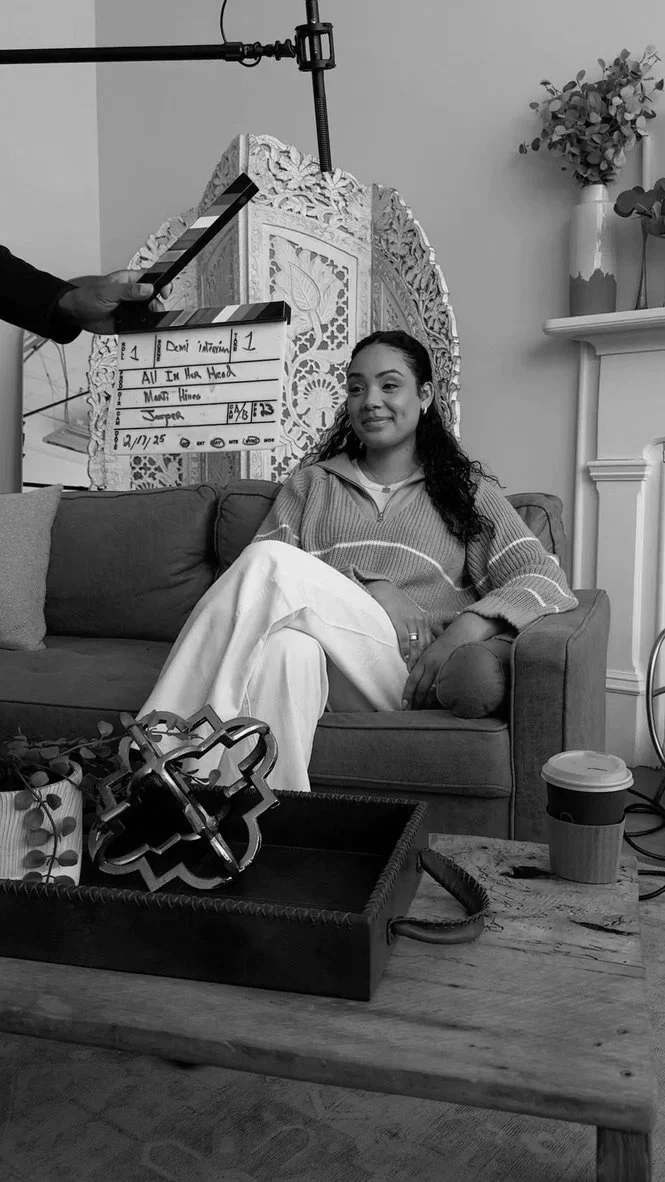 Behind-the-scenes photo of Marti Hines sitting on a sofa during a film shoot, with a clapperboard and crew equipment visible.