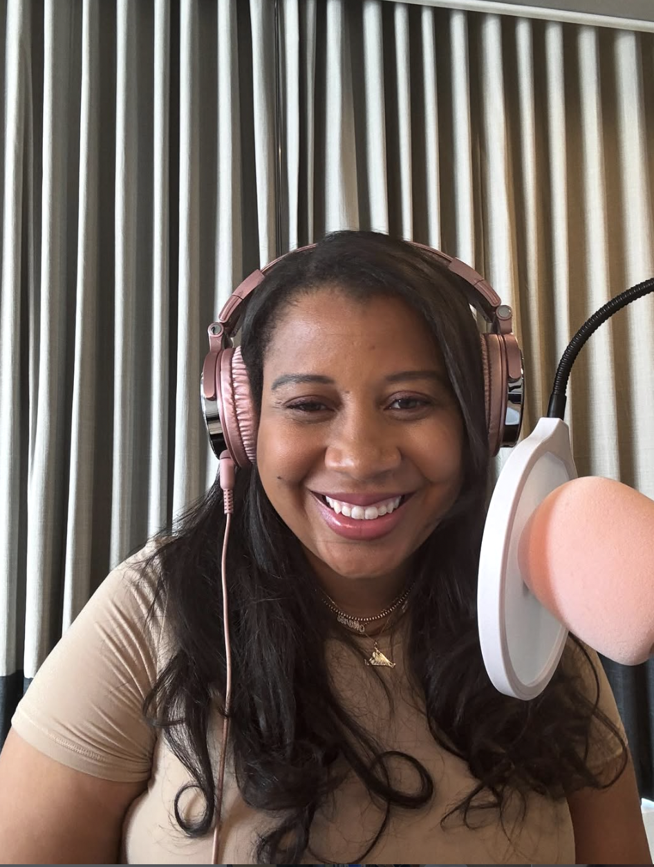 Marti Hines, a woman, smiling woman wearing pink headphones and a beige top, speaking into a microphone in a recording studio with beige curtains in the background.