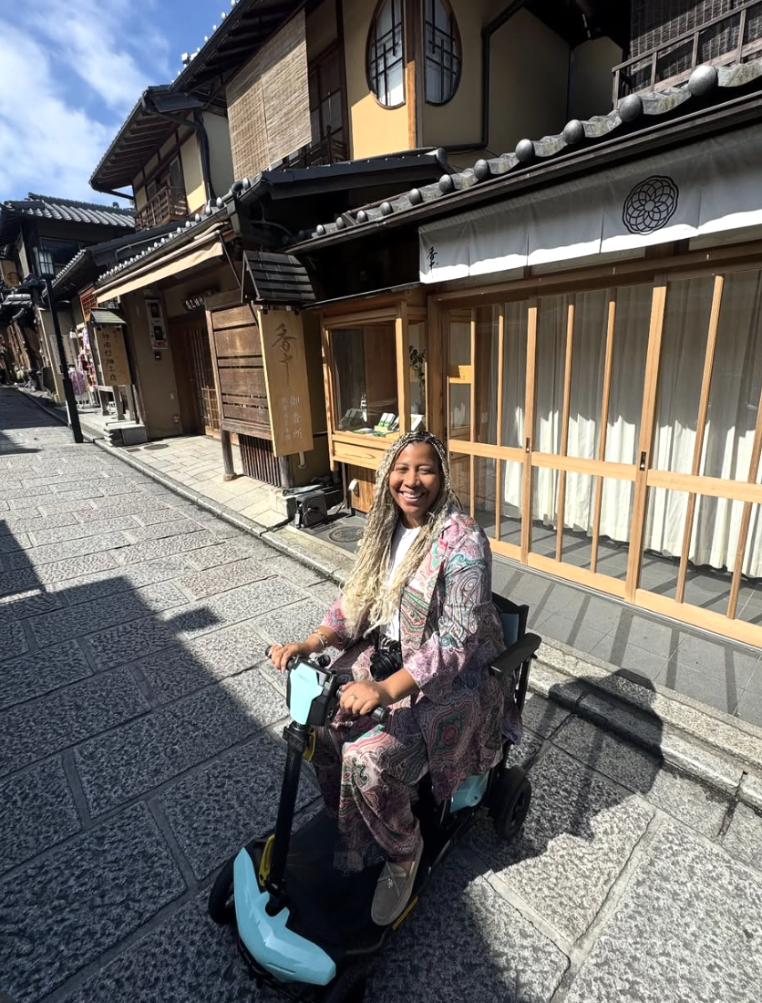 Smiling Marti Hines, a woman with long braids in a patterned outfit riding a light blue electric scooter on a stone-paved street with traditional Japanese buildings in the background.
