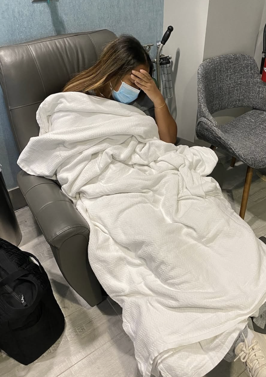 Marti Hines, a woman sitting on a hospital bed, wearing a blue face mask, with her head resting on her hand, getting treated for her Multiple Sclerosis, covered with white blankets, and wearing white sneakers.
