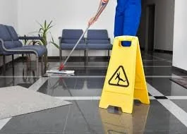 A person mopping a wet floor in a building hallway with a yellow caution sign.