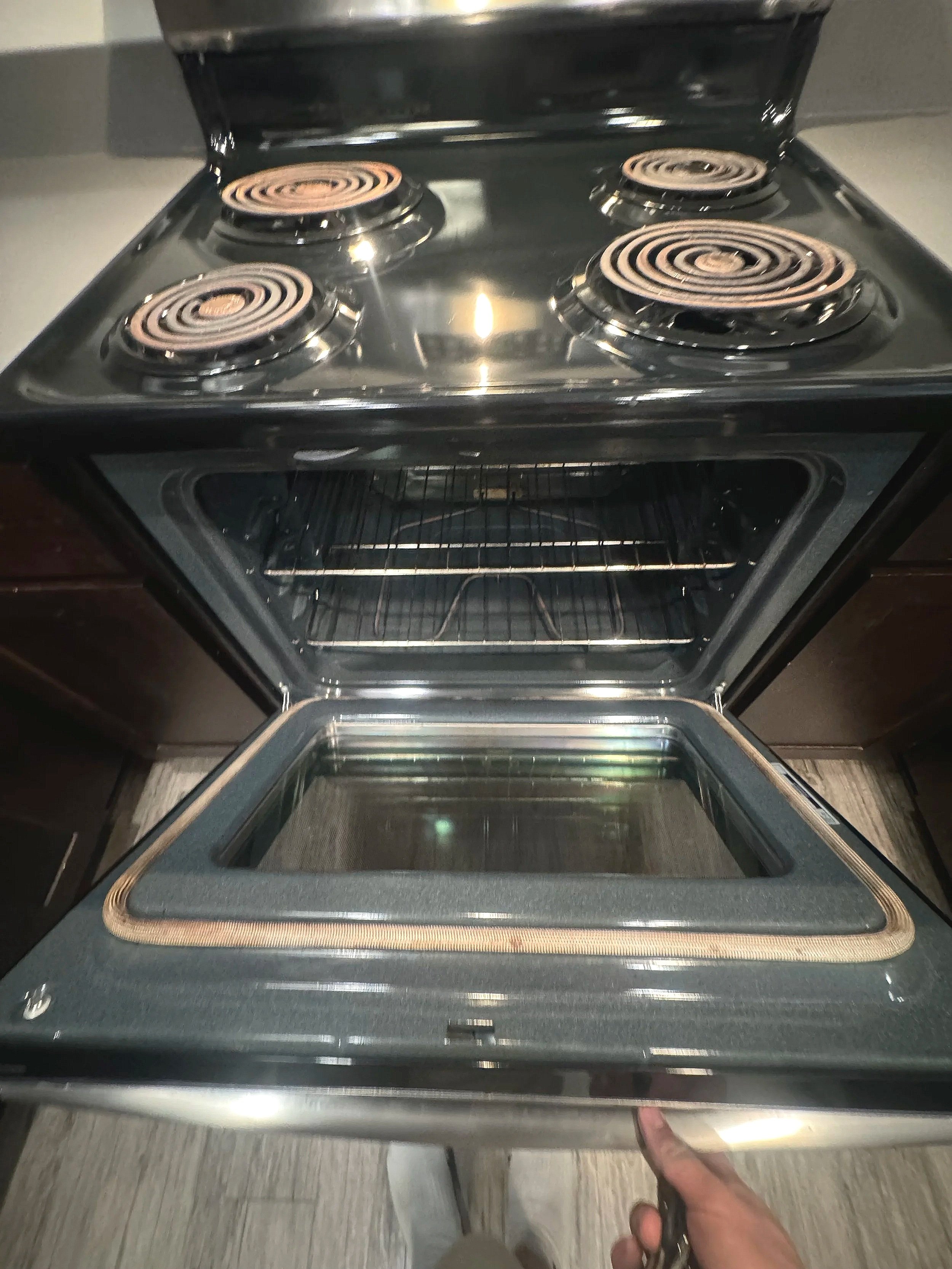 Open stainless steel oven with electric stovetop. Four coil burners on stovetop, oven door with glass window, and interior racks visible.