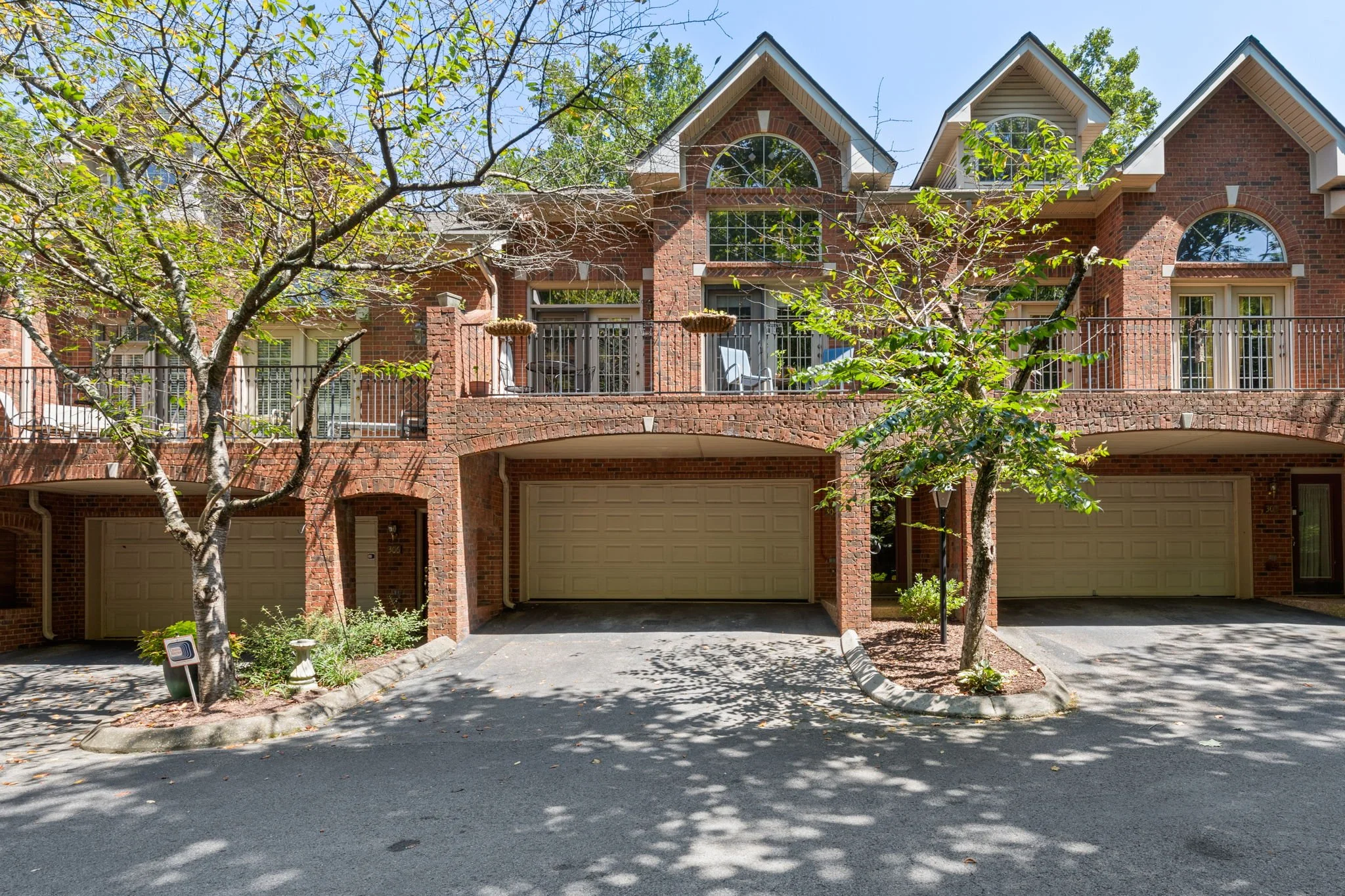 Front view of a red brick townhouse with two attached garages, trees with green leaves, and a balcony with patio furniture.