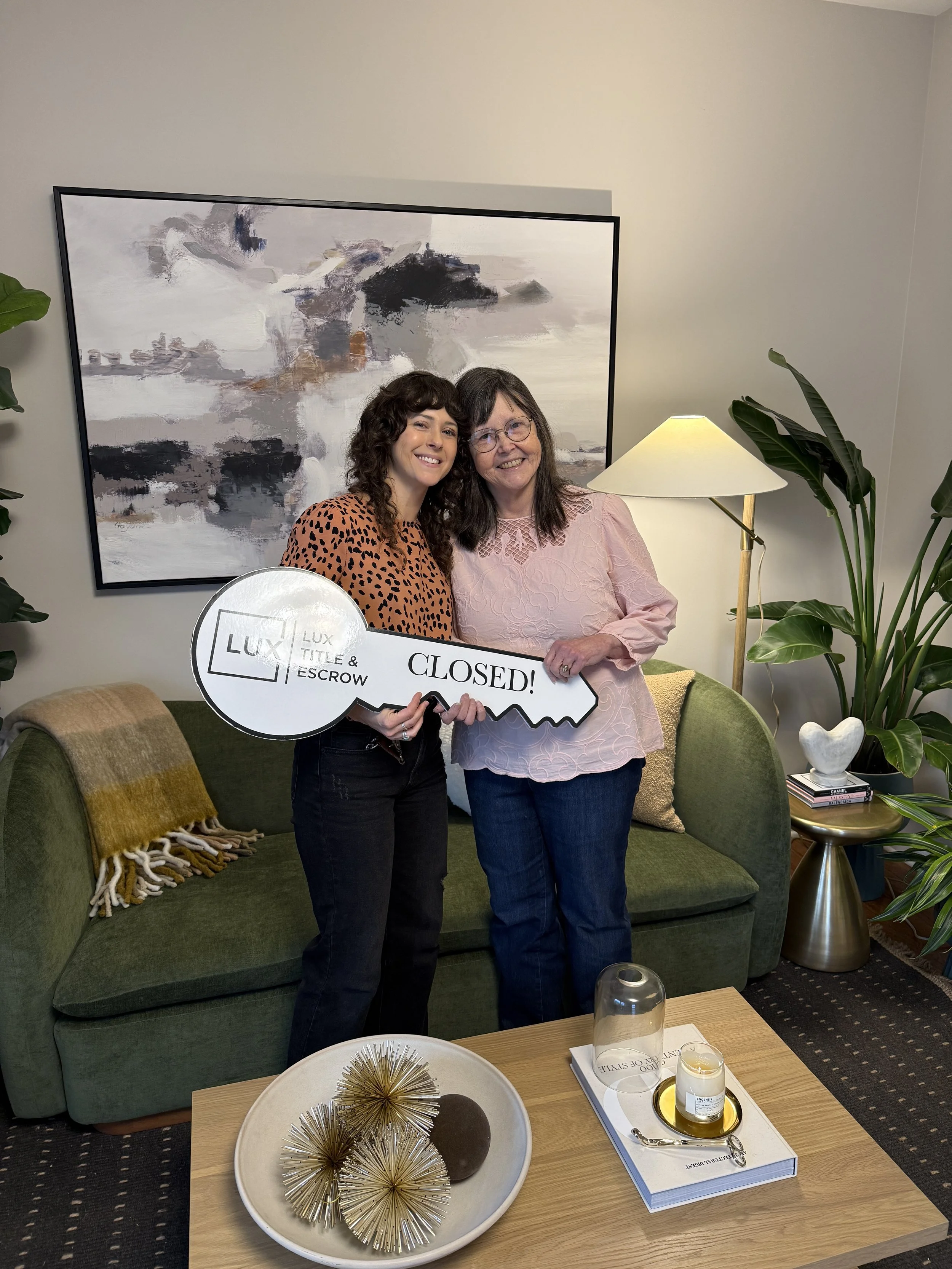 Two women standing together on a green couch, smiling and holding a large key-shaped sign that says "CLOSED!" in a cozy, well-decorated living room. One woman has curly dark hair and is wearing a leopard print top; the other has straight dark hair, g