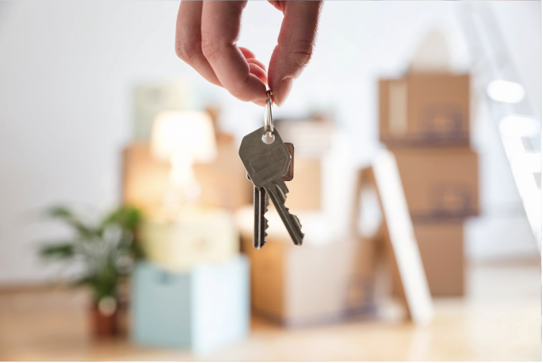 Person holding a key with two keys attached, with moving boxes in the background.