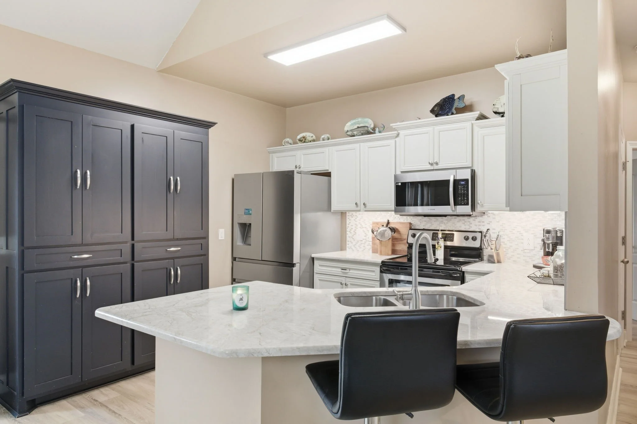 Modern kitchen with white cabinets, stainless steel refrigerator, microwave, oven, and a marble island with two black chairs.