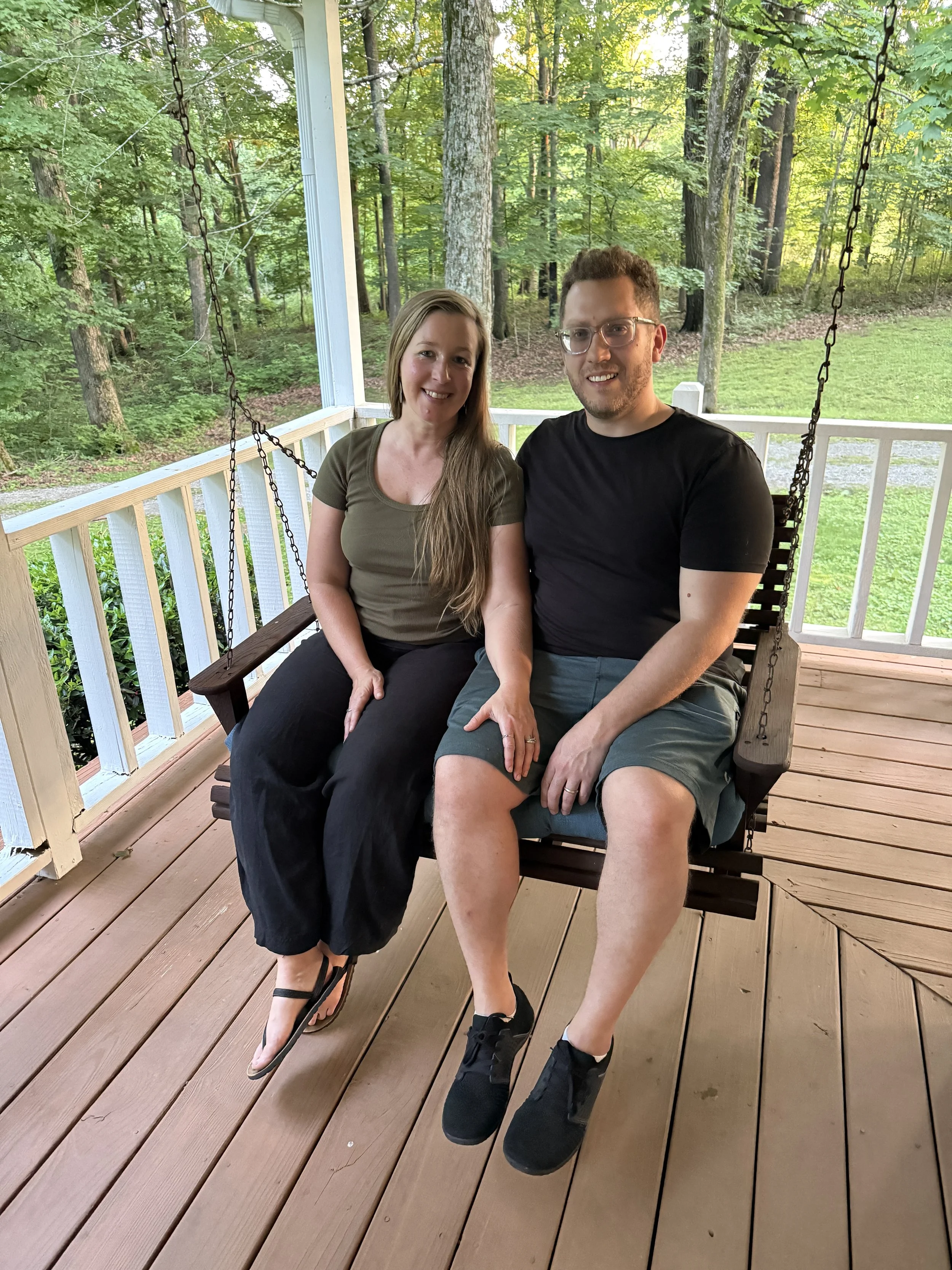 A smiling woman with long light brown hair and a man with short curly hair and glasses sitting on a porch swing, with a lush green forest background.