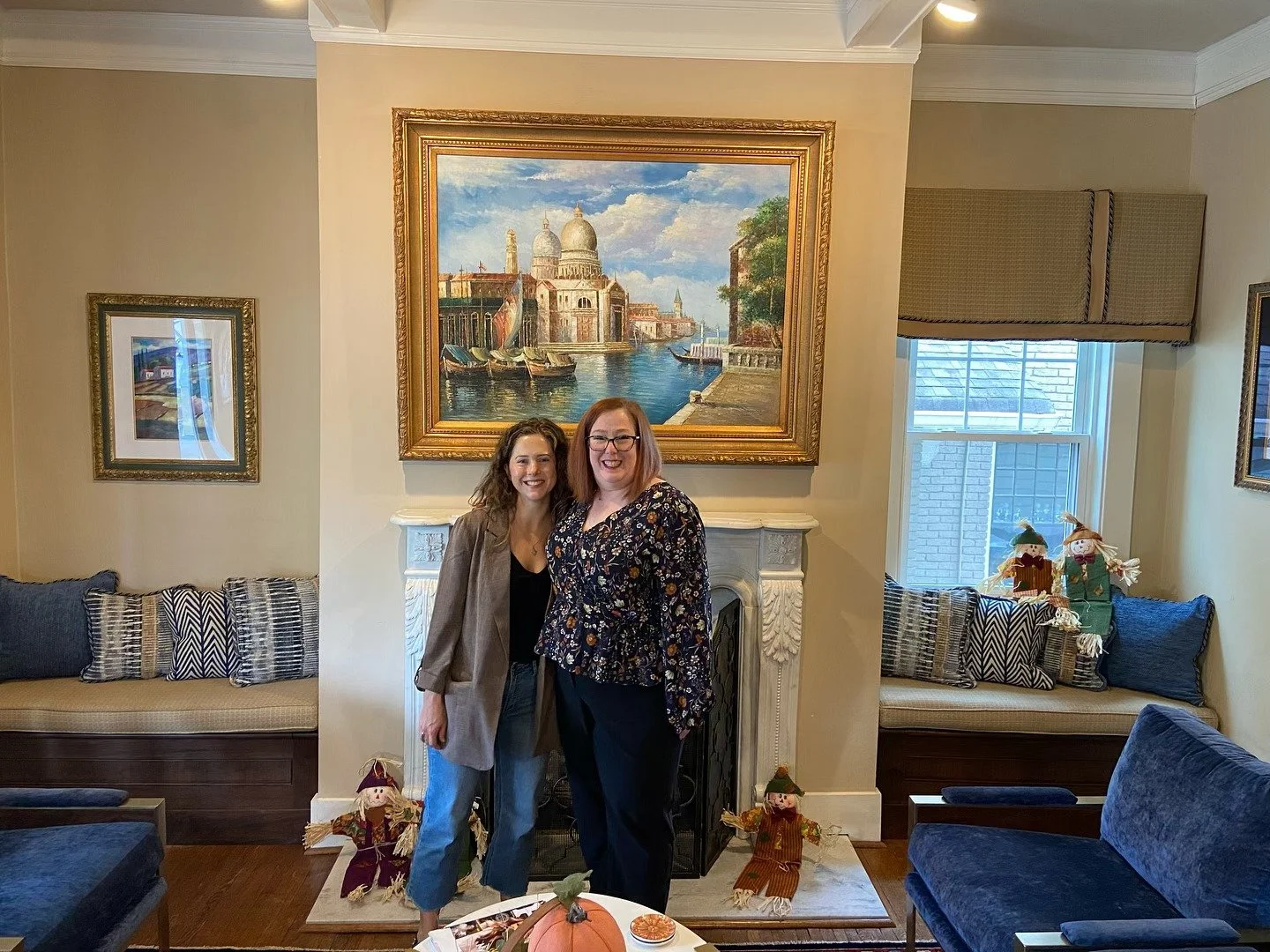 Two women standing in front of a fireplace in a living room, smiling. One woman has curly brown hair and is wearing a gray blazer, the other has straight red hair and is wearing glasses and a floral top. The fireplace has a large framed painting of V