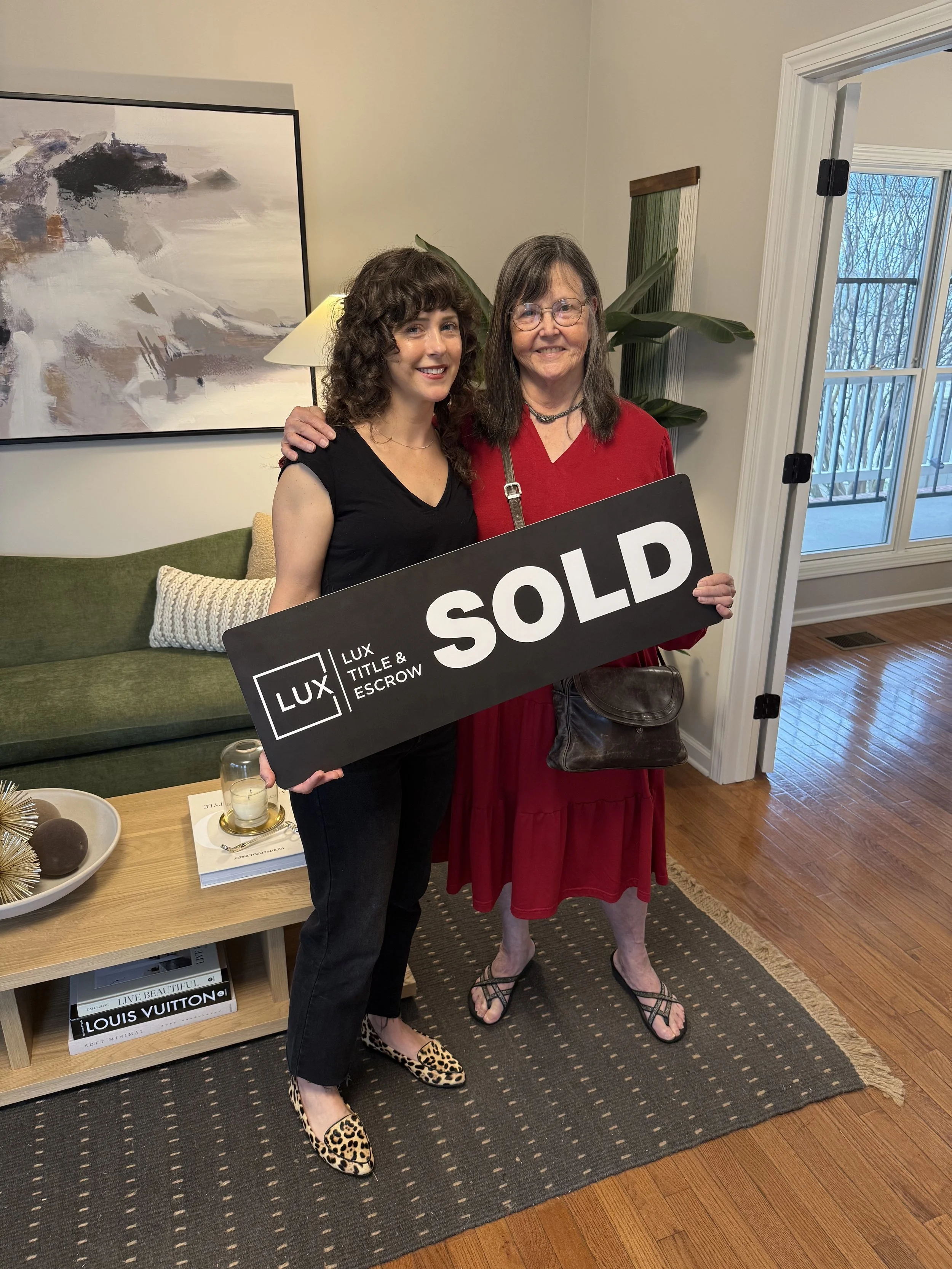 Two women standing in a living room holding a large black sign that reads 'SOLD' with the logo of Lux Title & Escrow. One woman is wearing a black top and leopard-print shoes, the other is wearing a red dress and sandals. They are smiling and standin
