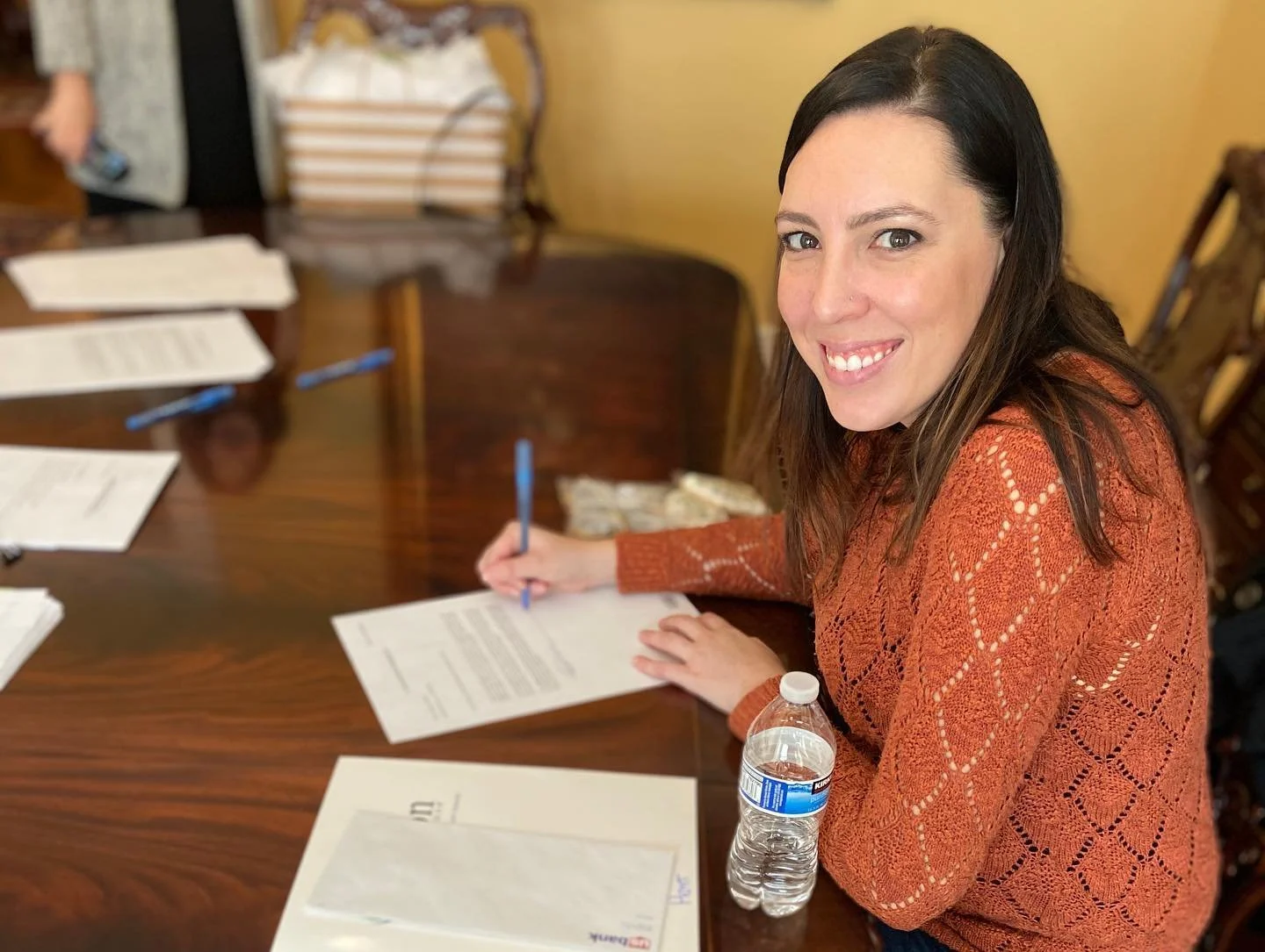 A woman sitting at a wooden table, smiling and signing a document with a blue pen. She has long brown hair and is wearing an orange knit sweater. There are papers, pens, and a water bottle on the table.