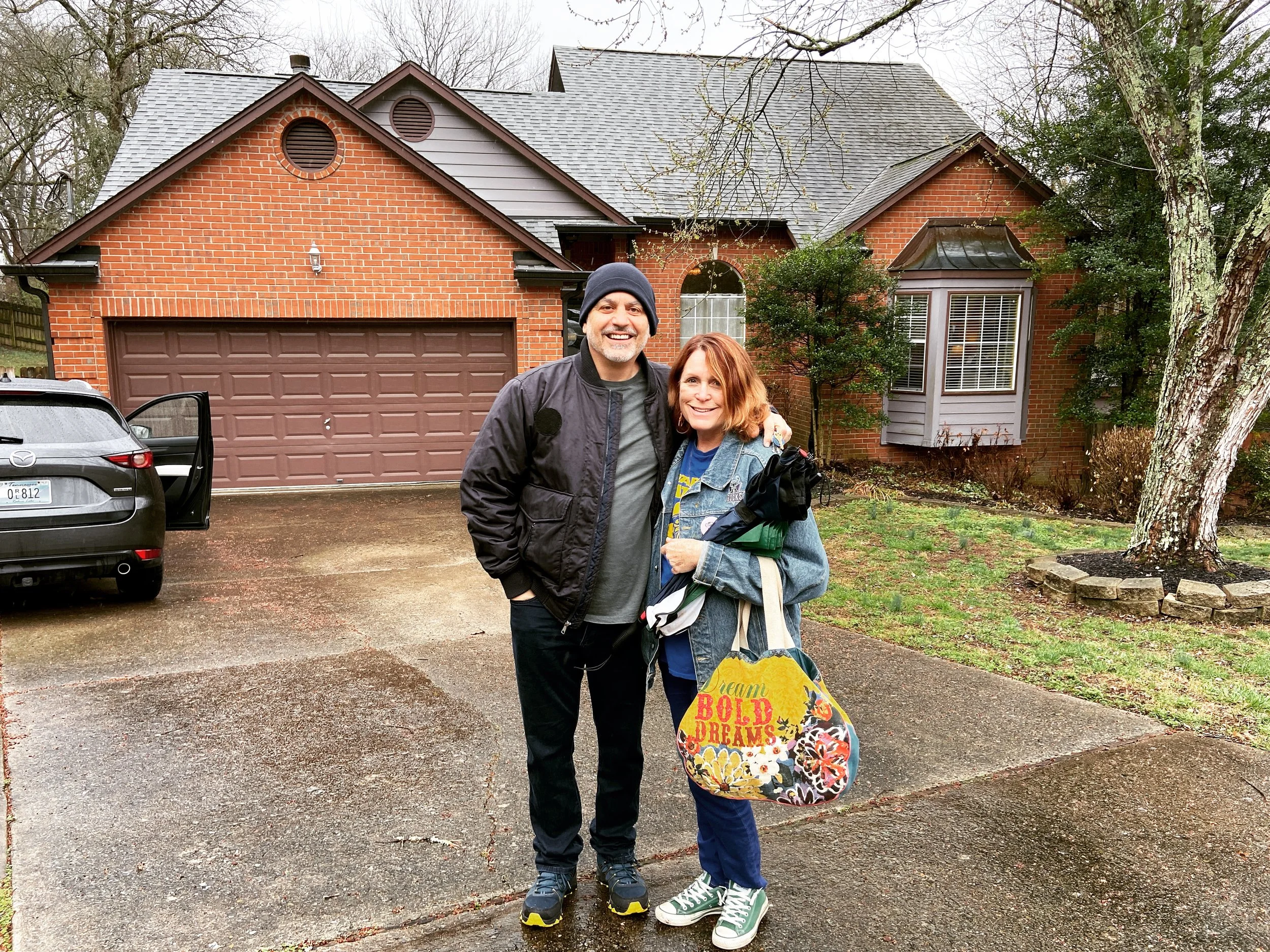 A man and woman standing outside a brick house on a rainy day, smiling and posing for the photo. The woman holds a colorful bag that says "Dream Bold Dreams." The man is wearing a black jacket and beanie, and the woman is wearing a denim jacket and g