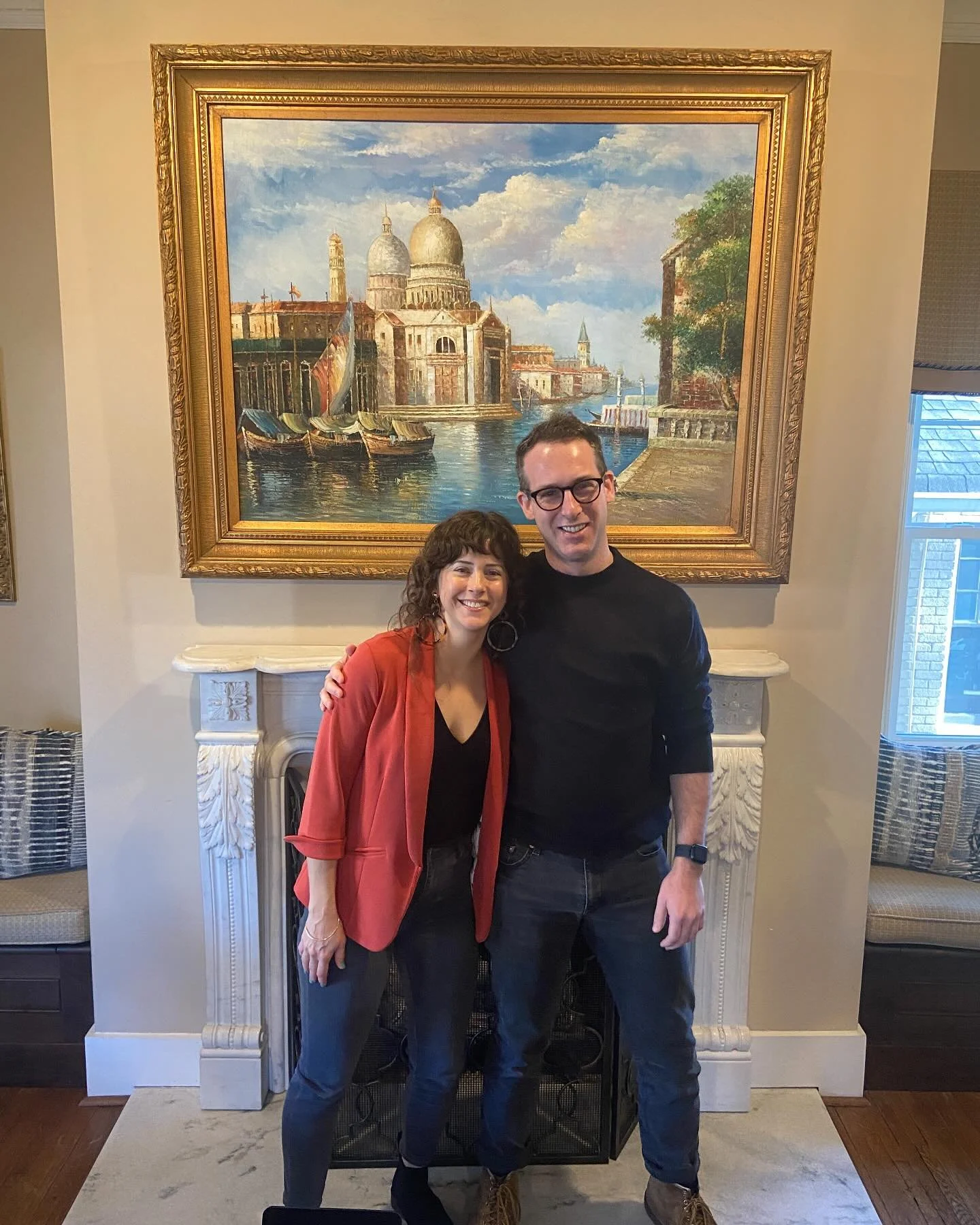 A smiling woman and man standing close together in front of the fireplace, with a large framed painting of Venice, Italy, hanging above them. The woman is wearing a red blazer over black and the man is dressed in black, with glasses and a watch.