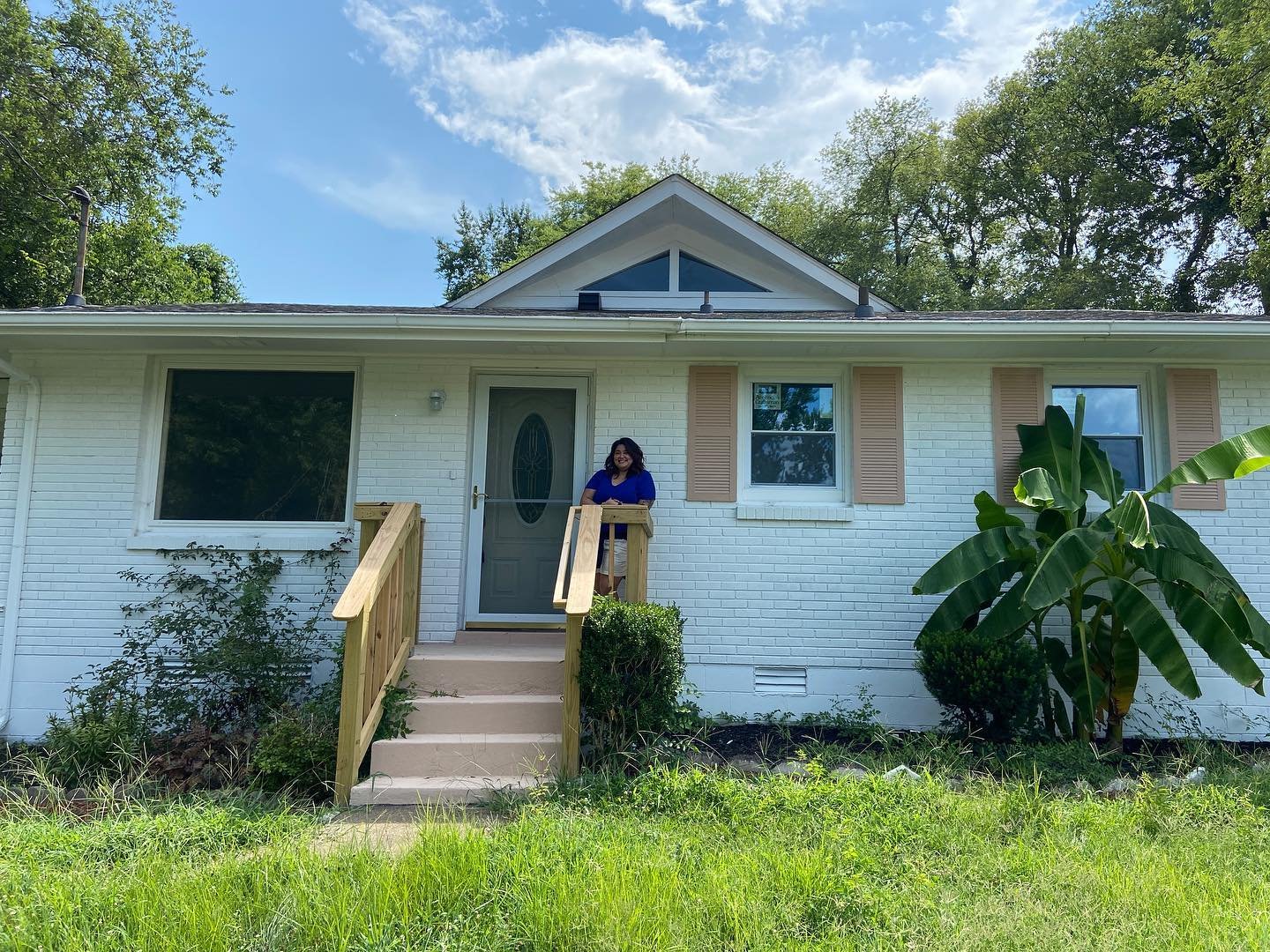 A woman standing on the steps of a white brick house with a green front door and two windows, surrounded by green trees and plants, under a partly cloudy blue sky.
