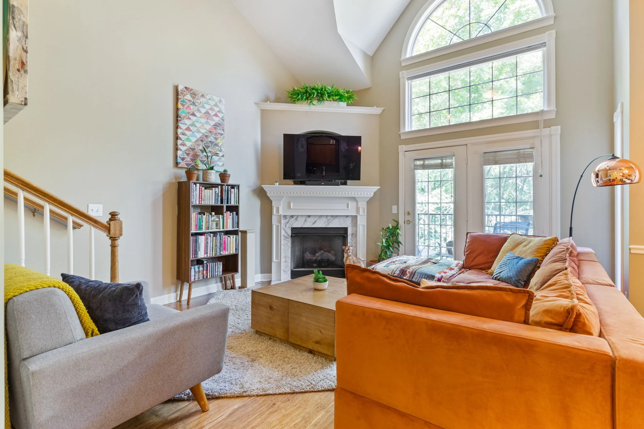 Living room with a fireplace, large windows, and a flat-screen TV above the fireplace. Orange sofa, gray armchair with pillows, wooden coffee table, bookshelf, and a floor lamp present.