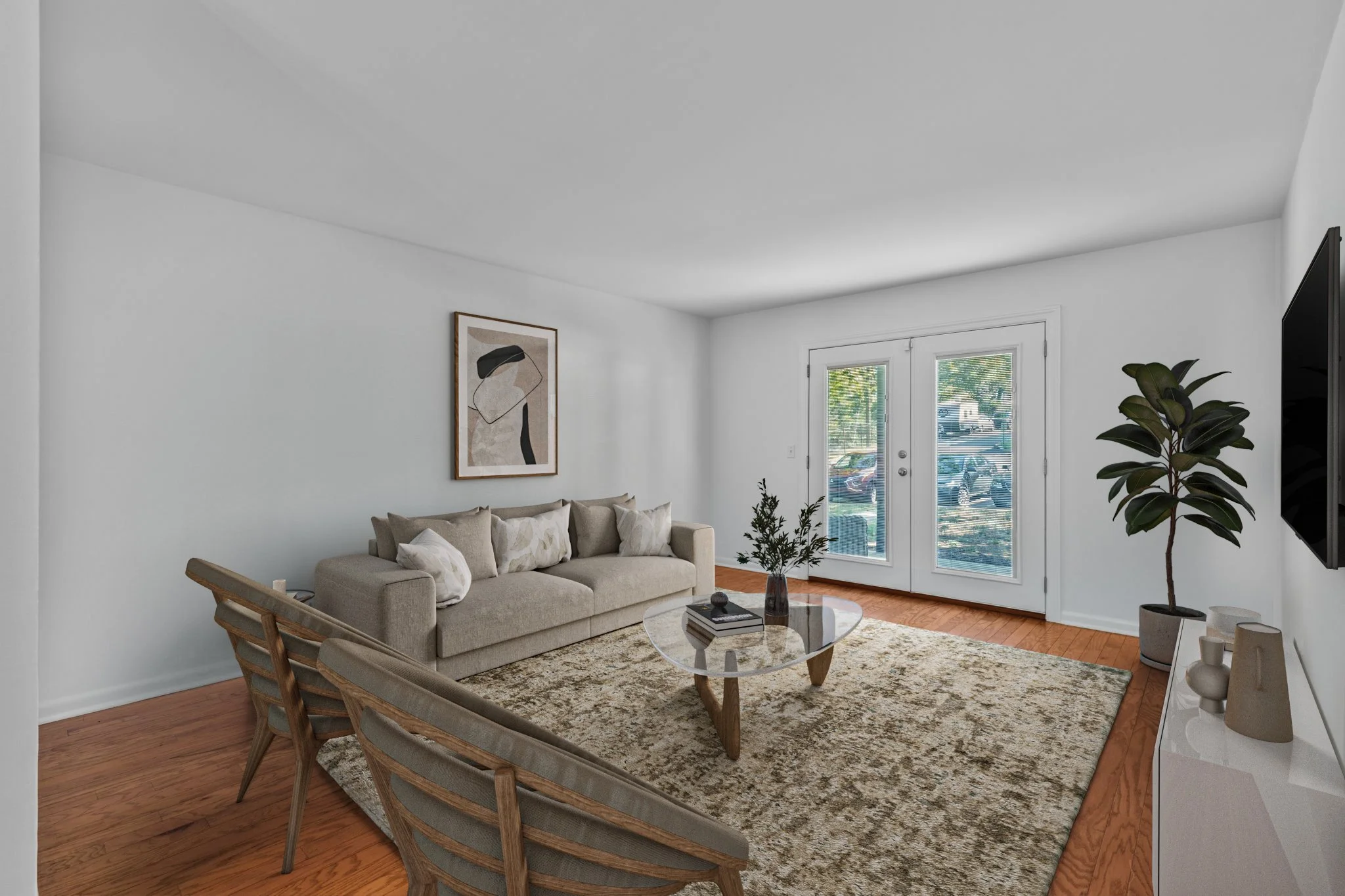 Living room with beige sofa, abstract artwork on white wall, glass coffee table with decor, beige rug, wooden chairs, potted plant, large glass doors, and a TV mounted on the wall.