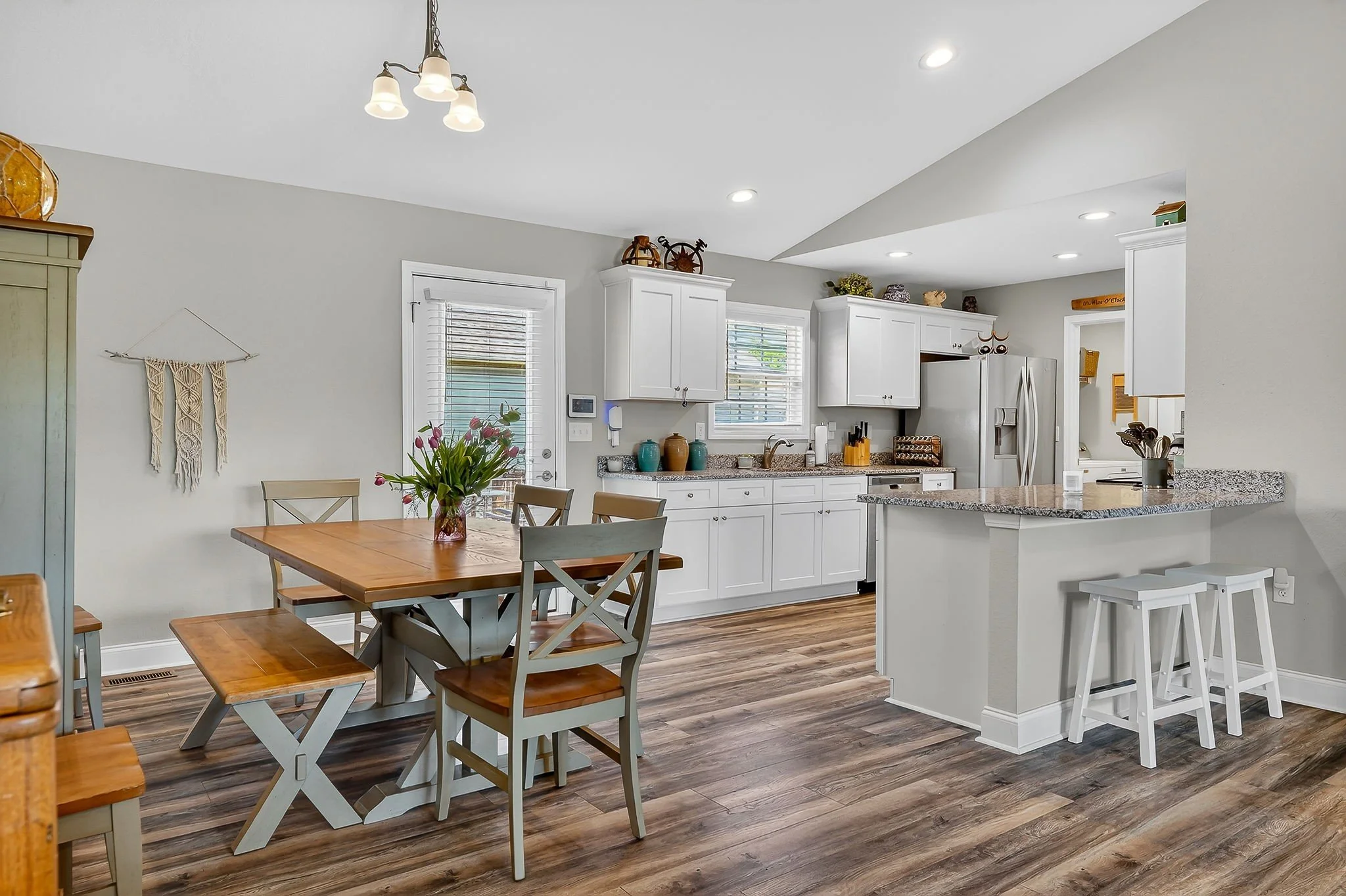 Open concept kitchen and dining area with white cabinets, stainless steel refrigerator, granite countertops, wooden dining table with chairs, and decorative items on top of cabinets. There are windows with blinds, wooden flooring, and ceiling lights.