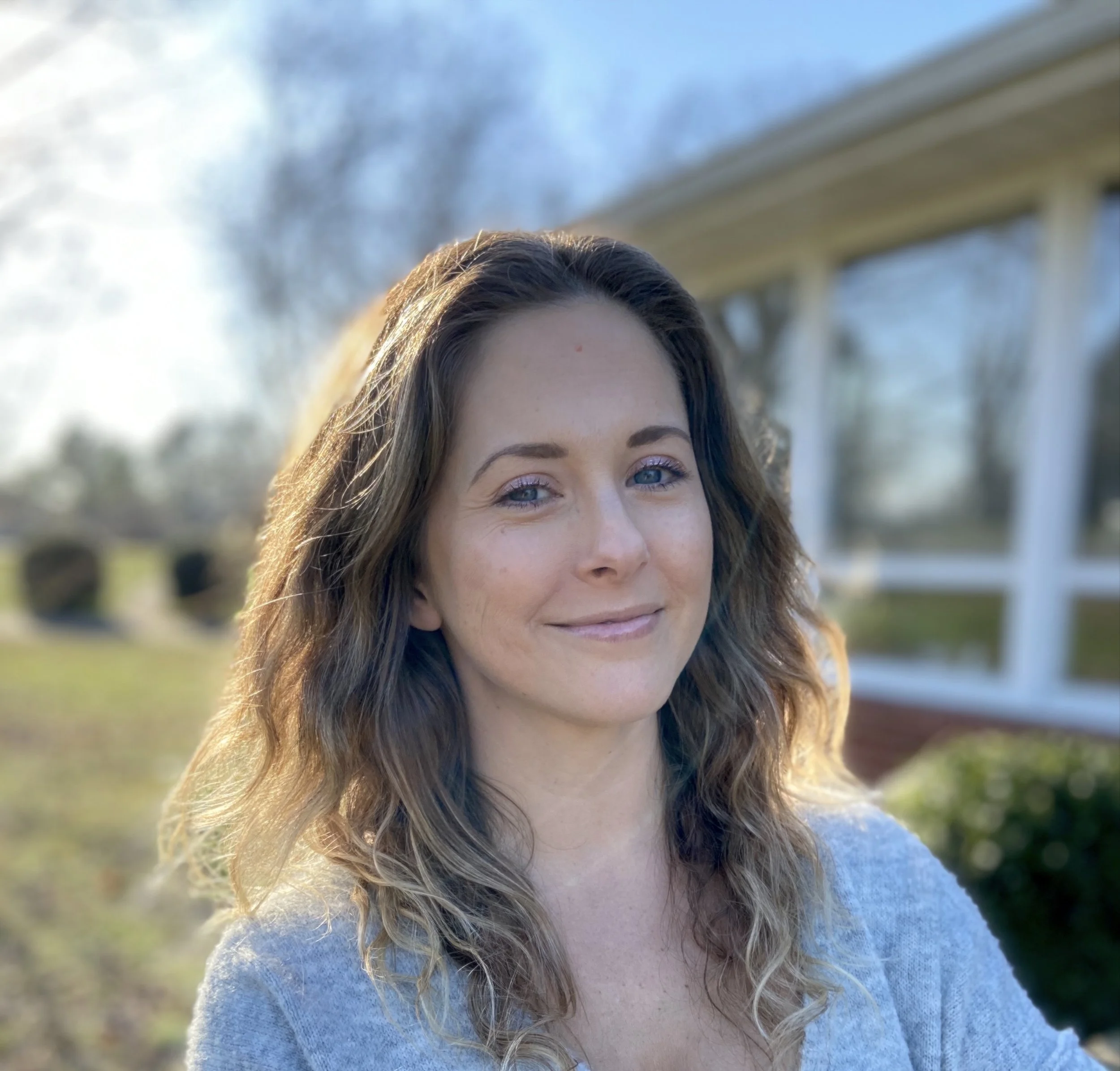 A woman with wavy brown hair and blue eyes smiling outdoors on a sunny day with a house and trees in the background.
