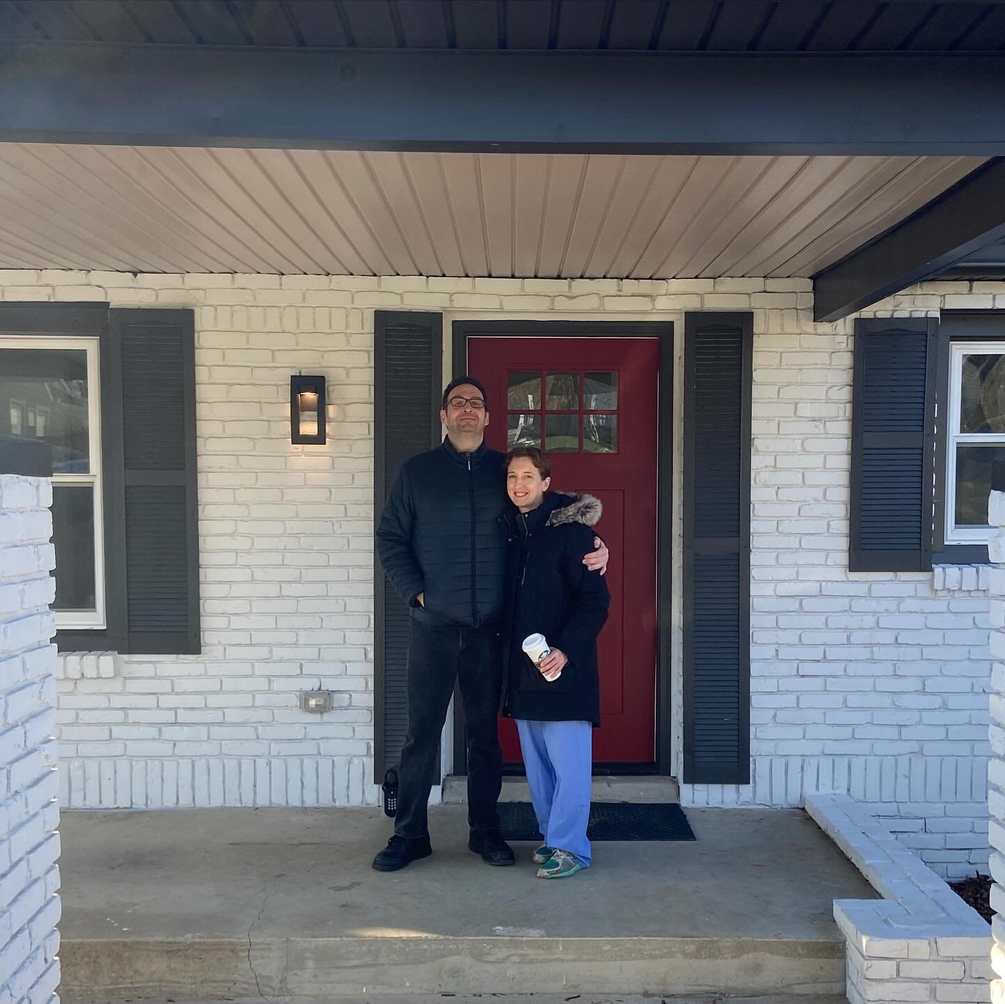 A man and a woman standing outside a white brick house with a red door and black shutters. The man is taller, wearing glasses and a black jacket, and the woman is smiling, holding a coffee cup, and wearing a black jacket with a fur-lined hood. They a