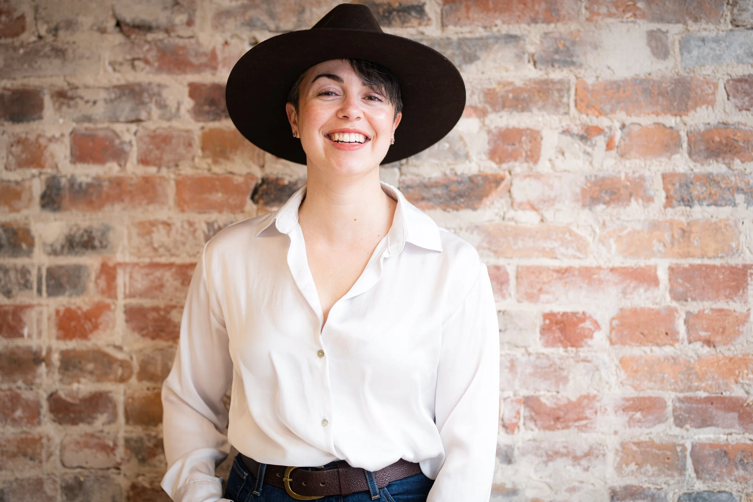 A woman wearing a white shirt and a wide-brimmed black hat, standing in front of an exposed brick wall, smiling.