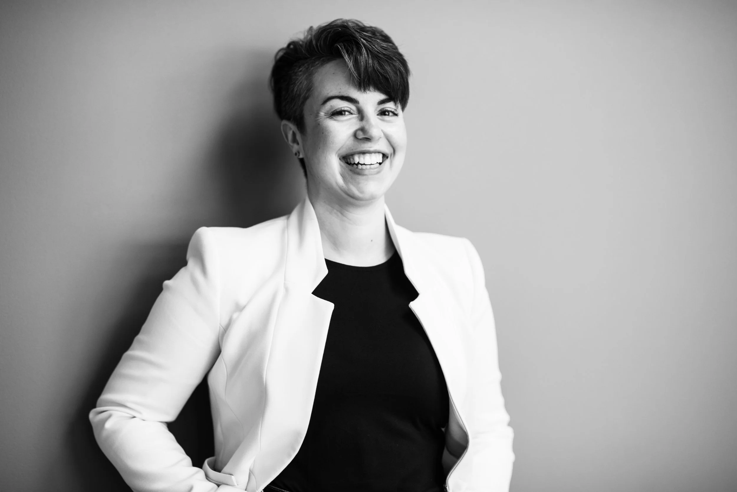 Black and white portrait of a woman with short hair, smiling, wearing a blazer and top, standing against a plain background.