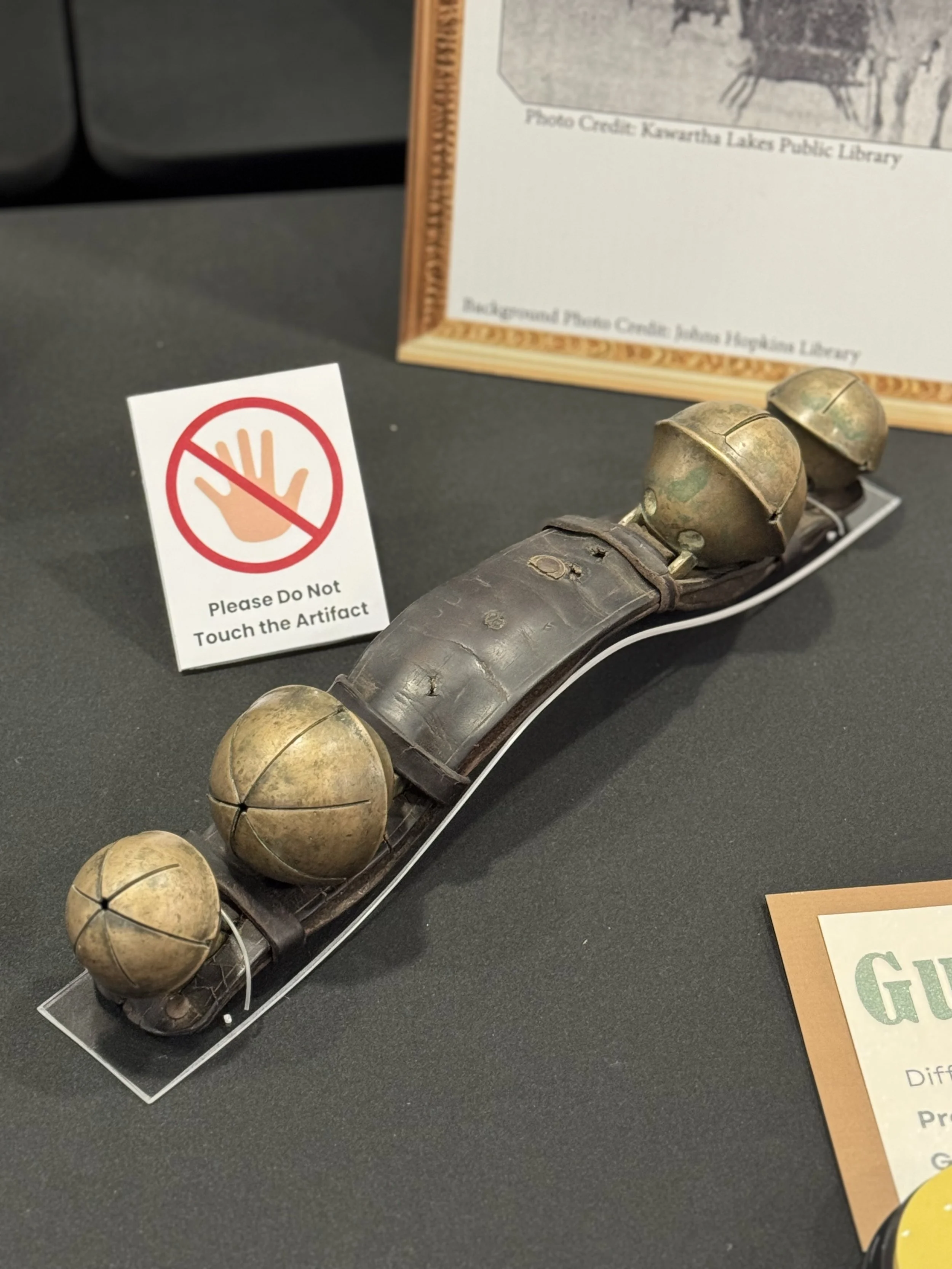 Antique sleigh bells on a plexiglass mount sitting on a black table cloth with a "Do Not Touch the Artifact" sign in the background.