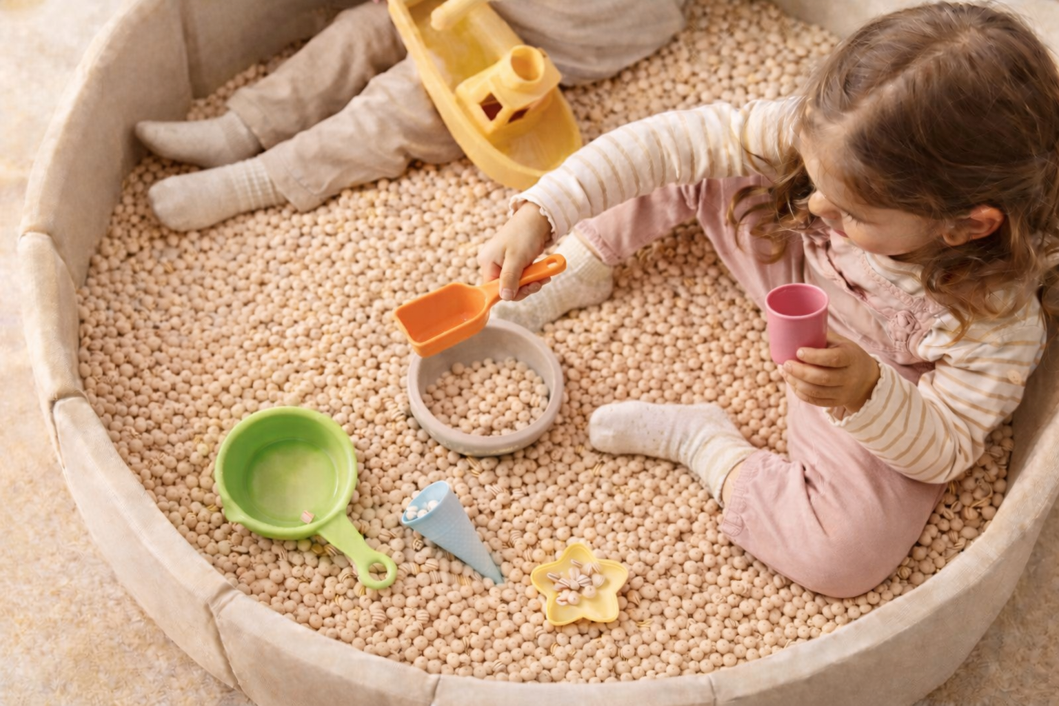 Ein Mädchen spielt in einem großen Sandkasten mit verschiedenen Spielsachen, darunter eine Schaufel, eine Tasse, eine Schüssel, eine Waffel-Form und ein kleines Boot.
