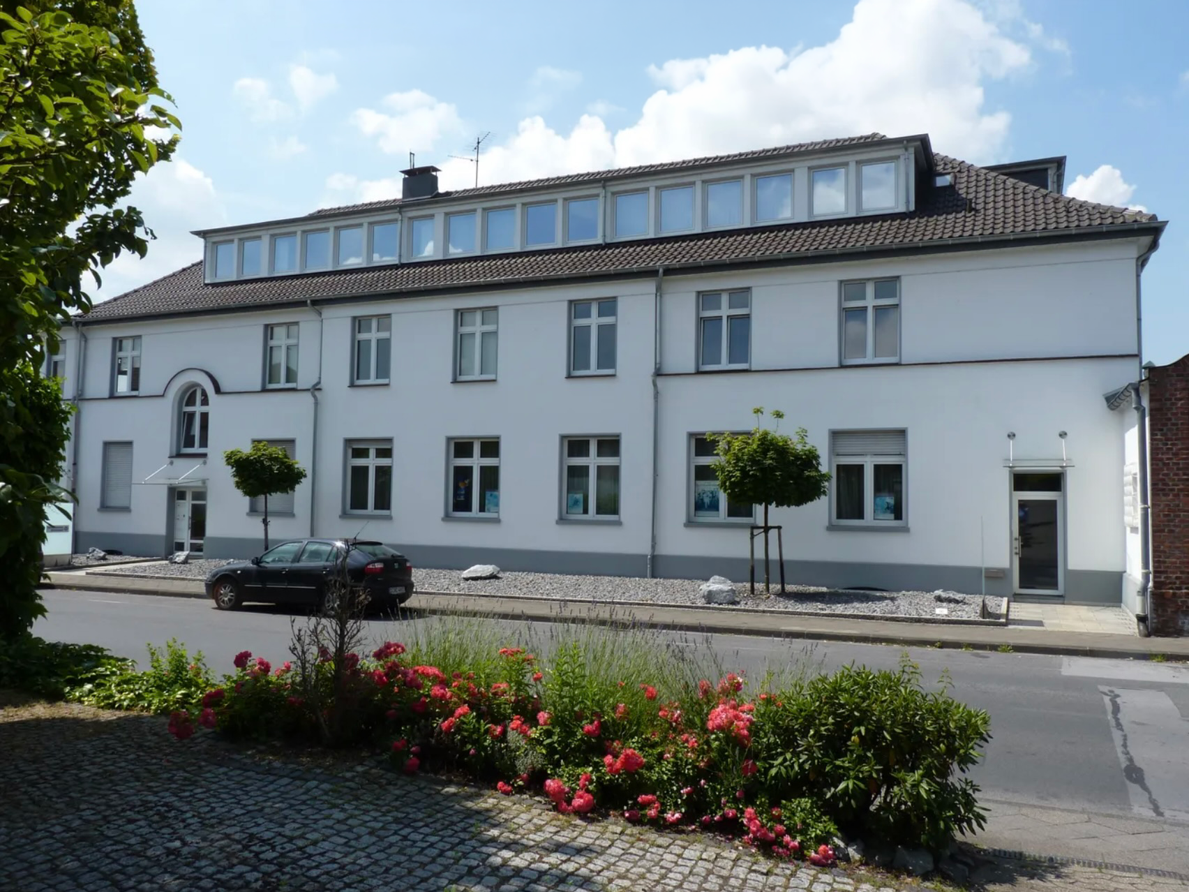 Außenansicht der Ergotherapiepraxis Daniela Engel. Weißes mehrstöckiges Gebäude mit mehreren Fenstern und Dachgauben, grüne Bäume und bunte Blumen im Vordergrund.